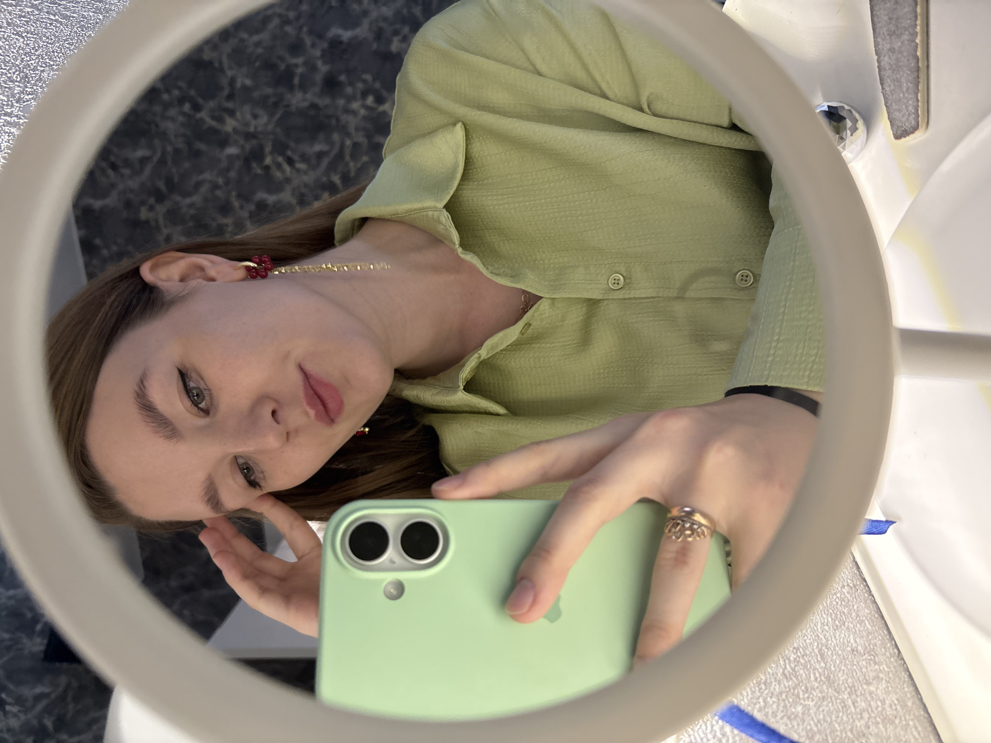 A young woman is reflected in a mirror, taking a selfie with her light green smartphone. She is wearing a light green collared shirt and has long, light brown hair. Her right hand is resting on her forehead, and she is wearing a gold ring on her left ring finger. She has a red dangle earring with gold accents in her left ear. The background of the reflection shows a marble-patterned wall. The mirror is circular with a white frame. The setting appears to be indoors, possibly a bathroom or dressing area, with a white countertop visible in the foreground.