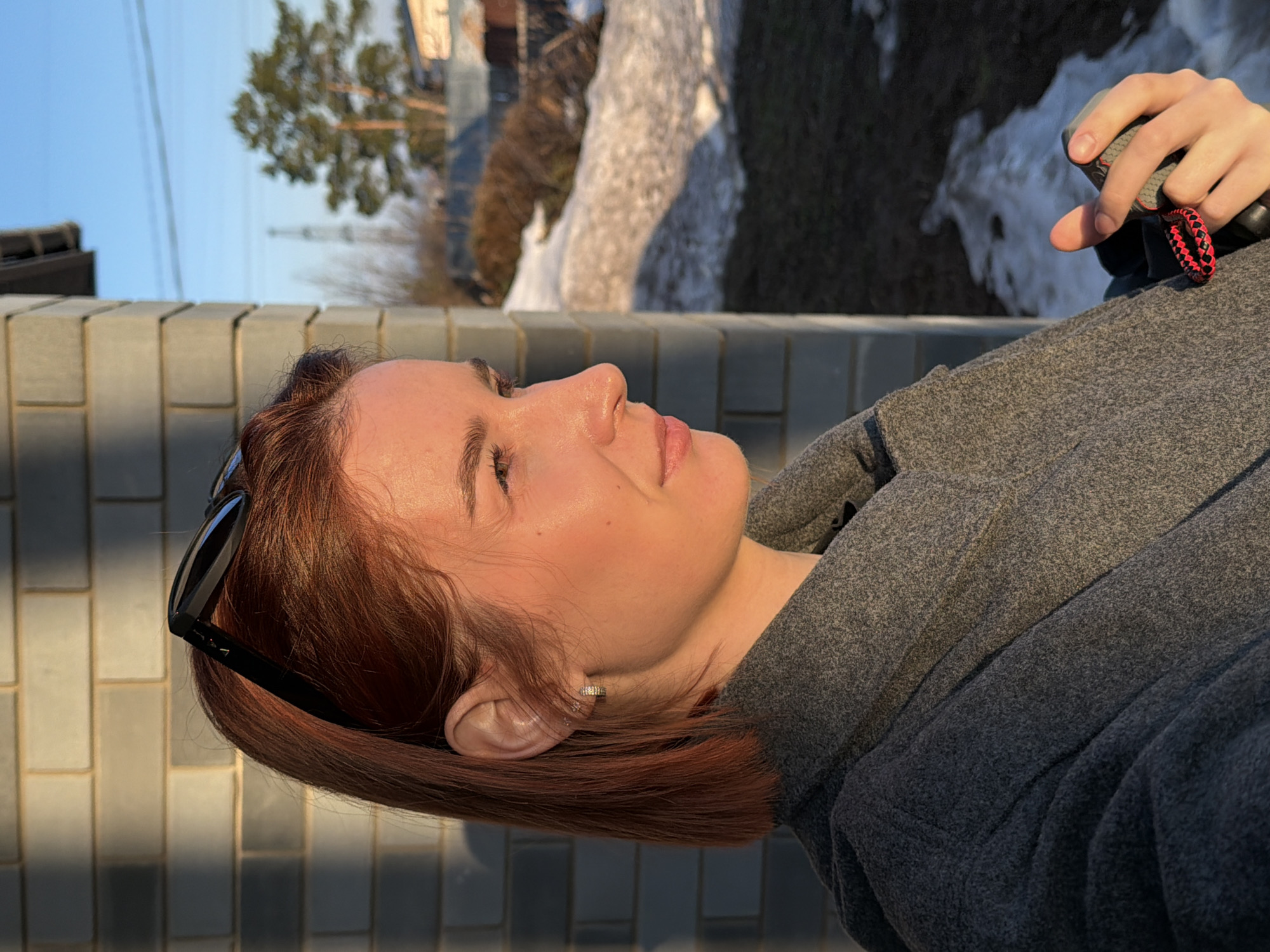 A young woman with reddish-brown hair is outdoors on a sunny day in what appears to be Chaikovsky, Russia, judging by the likely location. She is wearing a gray hooded jacket. Sunglasses are perched on her head. She is in profile. The background shows a fence, a tree, patches of snow, and buildings. She is holding something in her hand.