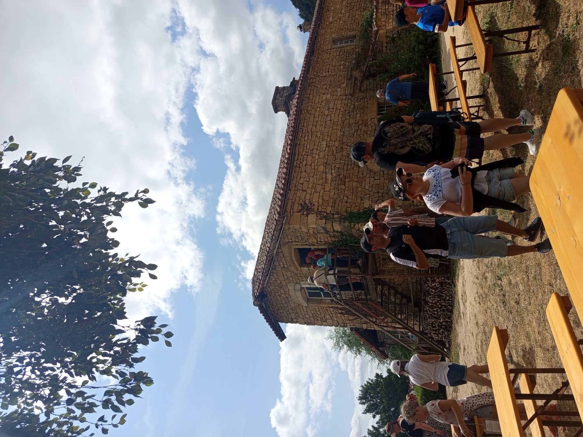 The image depicts an outdoor scene at the Azannes-et-Soumazannes, France historical site on a sunny day. A group of people are gathered around a stone building with a red tile roof. Several individuals are on a wooden staircase leading up to the building's entrance, while others stand on the ground. In the foreground, wooden picnic tables are set up on a dry, grassy area. The sky is blue with scattered clouds, and trees with green leaves are visible in the background, with one prominent tree branch with leaves in the upper left corner.