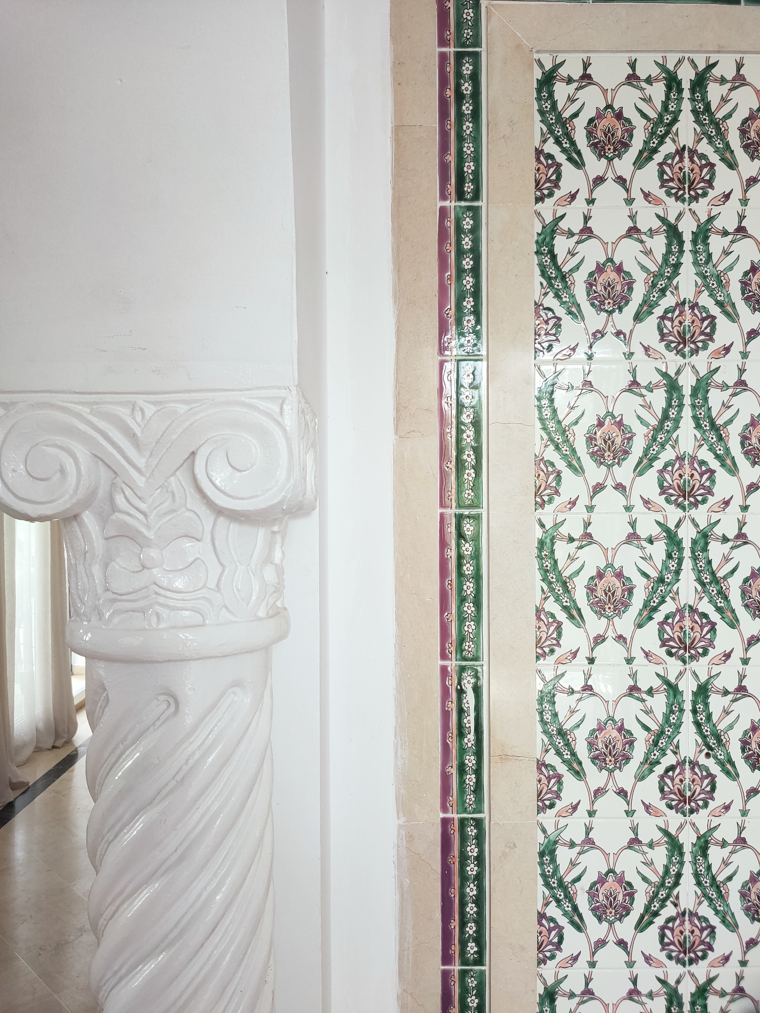 The image captures a detailed view of architectural elements, likely from an interior space, with a prominent column on the left and a tiled wall on the right. The column is white, with a decorative capital featuring volutes and intricate carvings, and a twisted shaft. The wall to the right is adorned with a repeating floral tile pattern in shades of green, pink, and white. This patterned section is bordered by a vertical strip of smaller tiles, alternating between purple and green with a delicate white floral motif. The background appears to be a plain white wall, with a subtle vertical seam. Hints of natural light suggest the presence of windows or an opening, with sheer curtains visible in the reflection on the column's surface. The overall impression is one of classical or ornate interior design. Based on the architectural style and tilework, this scene could be located in Mrezga, Tunisia, a region known for its historical and cultural influences reflected in its architecture.