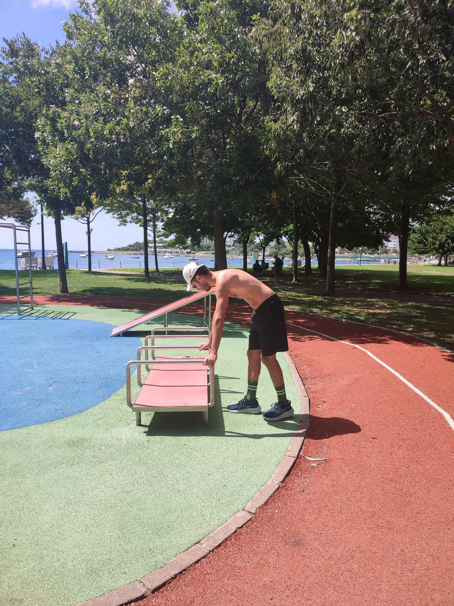 A shirtless, bare-chested man wearing black shorts, a white baseball cap, and dark blue athletic shoes with green socks is leaning over a pink and silver exercise bench in a park. The man is positioned on a red track surface with a white line. To his left, there is a green turf area, and further left, a blue surface. Behind the man, a body of water with boats is visible, with trees and buildings lining the distant shore. The scene appears to be a public park with outdoor exercise equipment, possibly located in Kadıköy, Turkey, given the style of the waterfront and architecture. The sky is blue with some clouds.