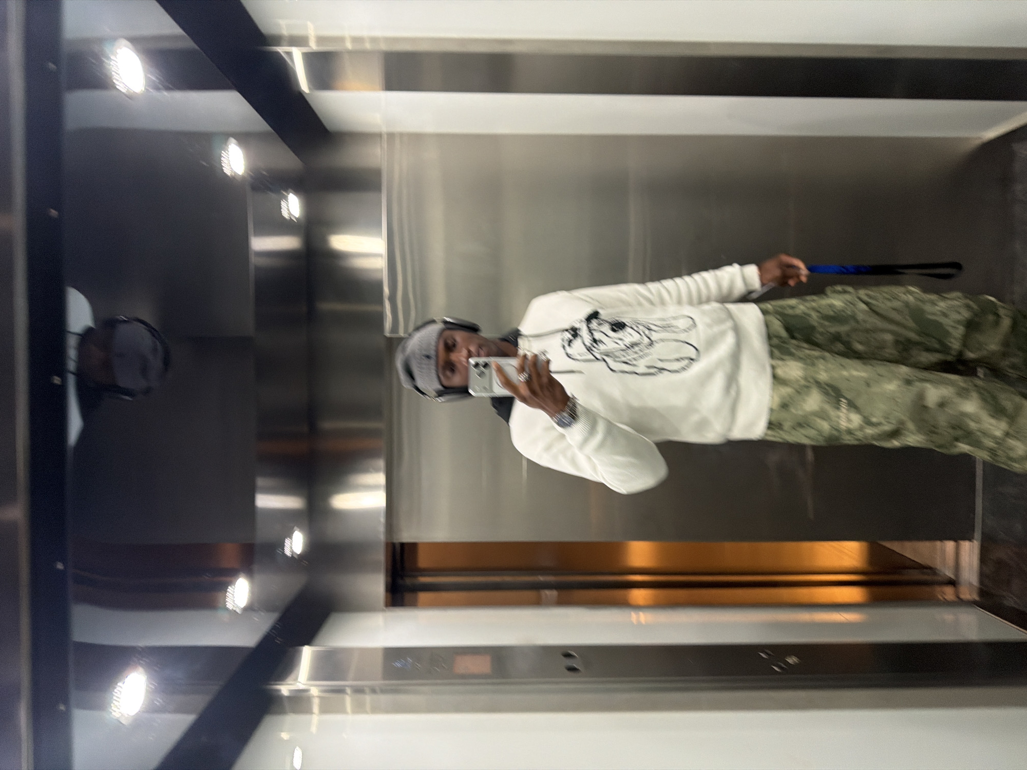 A person wearing a gray knit hat and a white long-sleeved shirt with a black graphic is standing in an elevator and taking a picture in the mirror with a cell phone. The person is also wearing camouflage pants. The elevator has stainless steel walls, a black ceiling with recessed lights, and a digital display panel. There are also a small stick-like object held between his fingers. The location could be an elevator in a building located in London, United Kingdom.