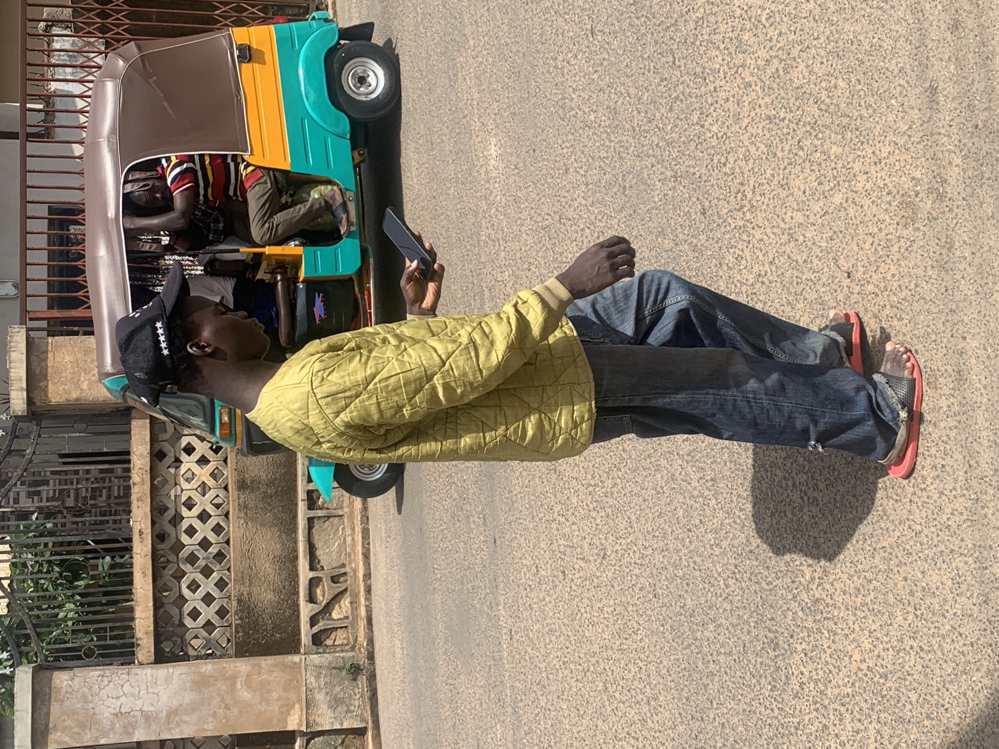 A man stands on a paved road in Barakin Akawo, Nigeria, looking at his phone. He is wearing jeans, a yellow quilted jacket, and red sandals. A colorful auto rickshaw is parked behind him, with two other people inside. A decorative wall and gate are to the left of the rickshaw.