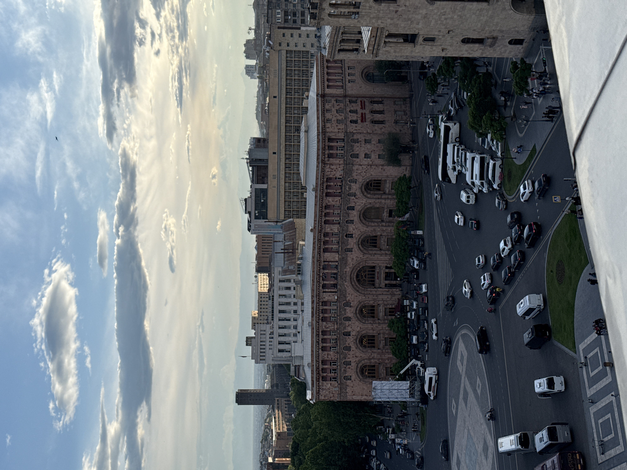 View of Yerevan, Armenia, including Republic Square. The sky above the buildings is partly cloudy. Below the buildings and the sky are an area with buildings, followed by a major road intersection and Republic Square with cars, buses, and people.