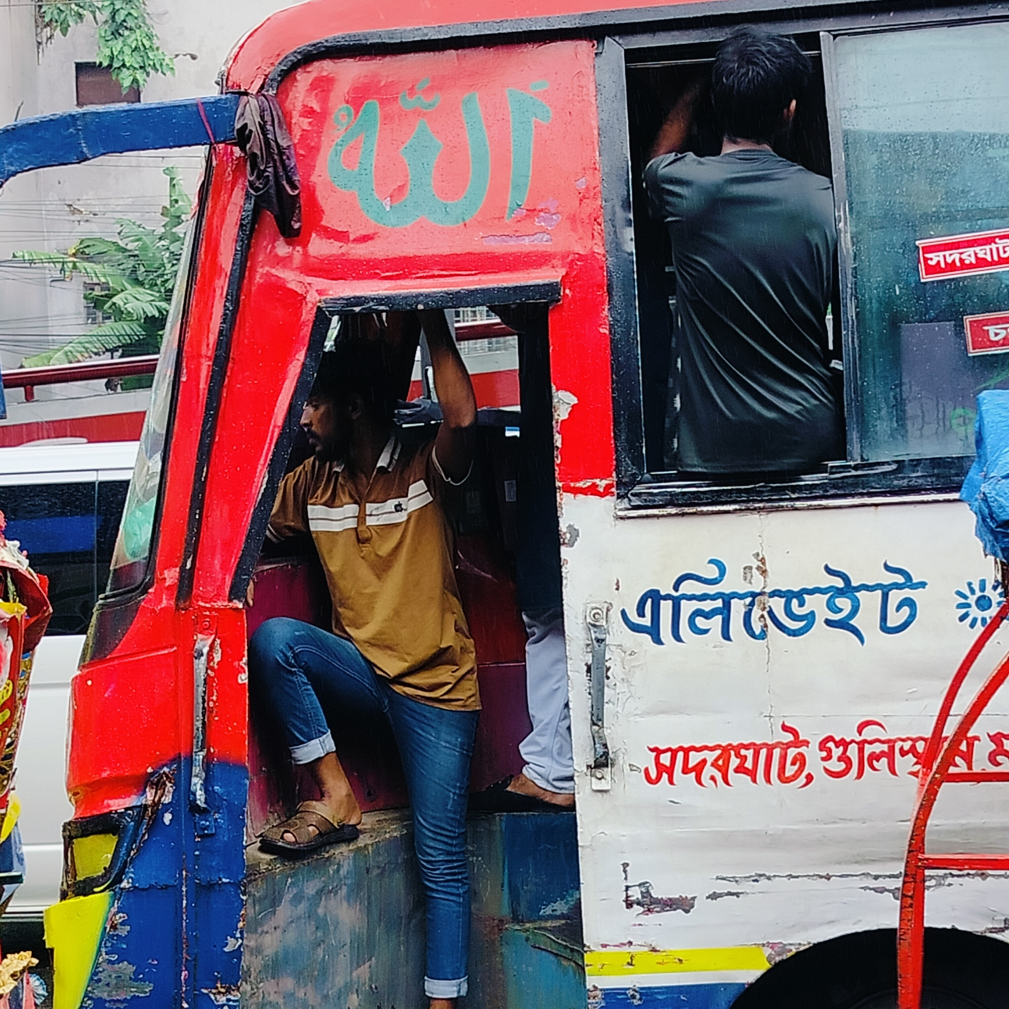 The image shows a red and blue bus, possibly a local bus in Dhaka, Bangladesh, with several people visible. A man in a brown and white striped polo shirt and blue jeans is sitting by the open door of the bus, with his legs extended outwards. He is wearing brown sandals. Another person, wearing a dark green t-shirt, is standing inside the bus near the window, with their back to the camera. The bus has some writing on its side in blue and red. On the front of the bus, there is writing in a teal color. The bus appears to be on a city street, with a white vehicle visible in the background. It is raining, as indicated by the wet surfaces and the texture of the image. The overall scene suggests a typical day on a busy street in Dhaka.