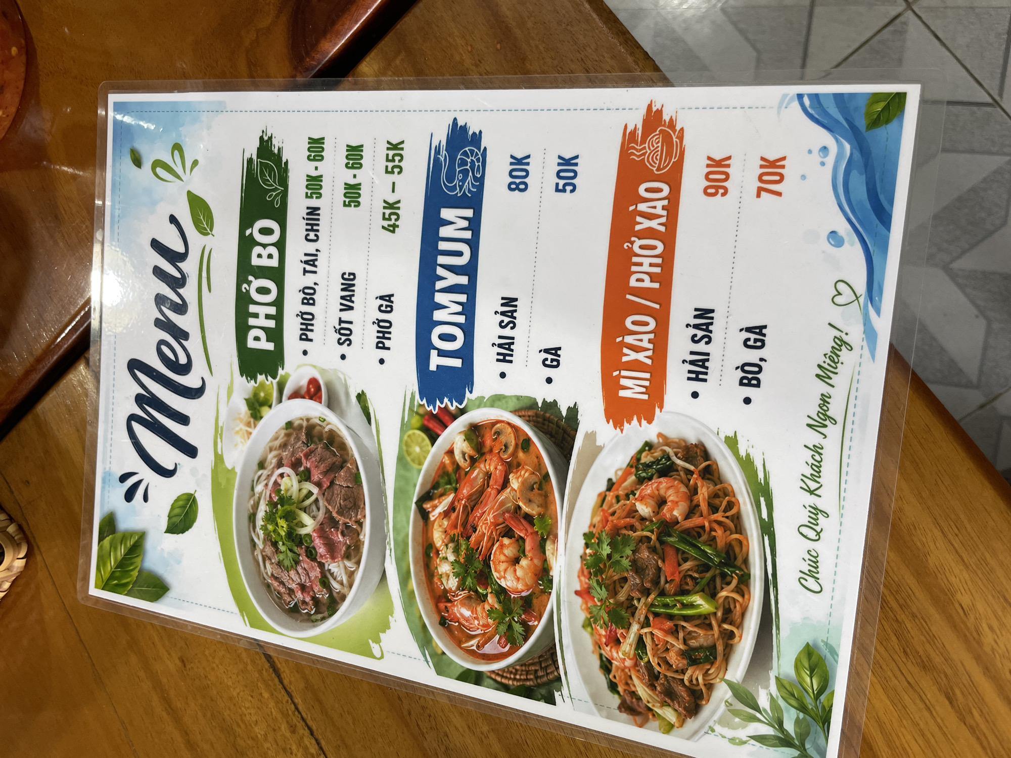A close-up, slightly angled shot captures a laminated Vietnamese restaurant menu resting on a warm-toned wooden table. The menu features a vibrant design with watercolor-style background elements and food illustrations.
The menu is divided into three main sections:
1. **PHỞ BÒ (Beef Pho)**: This section lists "PHỞ BÒ, TÁI, CHÍN" (Beef Pho with rare and well-done beef) priced at 50K - 60K VND, "SỐT VANG" (Red wine beef stew pho) also at 50K - 60K VND, and "PHỞ GÀ" (Chicken Pho) at 45K - 55K VND. An image of a steaming bowl of pho with sliced beef, noodles, and fresh herbs is displayed next to this section.
2. **TOMYUM**: This section offers "HẢI SẢN" (Seafood) Tom Yum for 80K VND and "GÀ" (Chicken) Tom Yum for 50K VND. A large, colorful bowl of Tom Yum soup with shrimp, mushrooms, and herbs is shown.
3. **MÌ XÀO / PHỞ XÀO (Stir-fried Noodles / Stir-fried Pho)**: This section includes "HẢI SẢN" (Seafood) for 90K VND and "BÒ, GÀ" (Beef, Chicken) for 70K VND. A plate of stir-fried noodles with shrimp, beef/chicken, and various vegetables is depicted.
The menu title "Menu" is prominently displayed in a decorative script. Below the listed items, the Vietnamese phrase "Chúc Quý Khách Ngon Miệng!" (Wish you a delicious meal!) is printed, accompanied by a small heart icon. The wooden table exhibits a visible grain pattern, and parts of the menu reflect light from the lamination. The setting appears to be a casual dining environment, likely in Phu Thuy Ward, Vietnam.