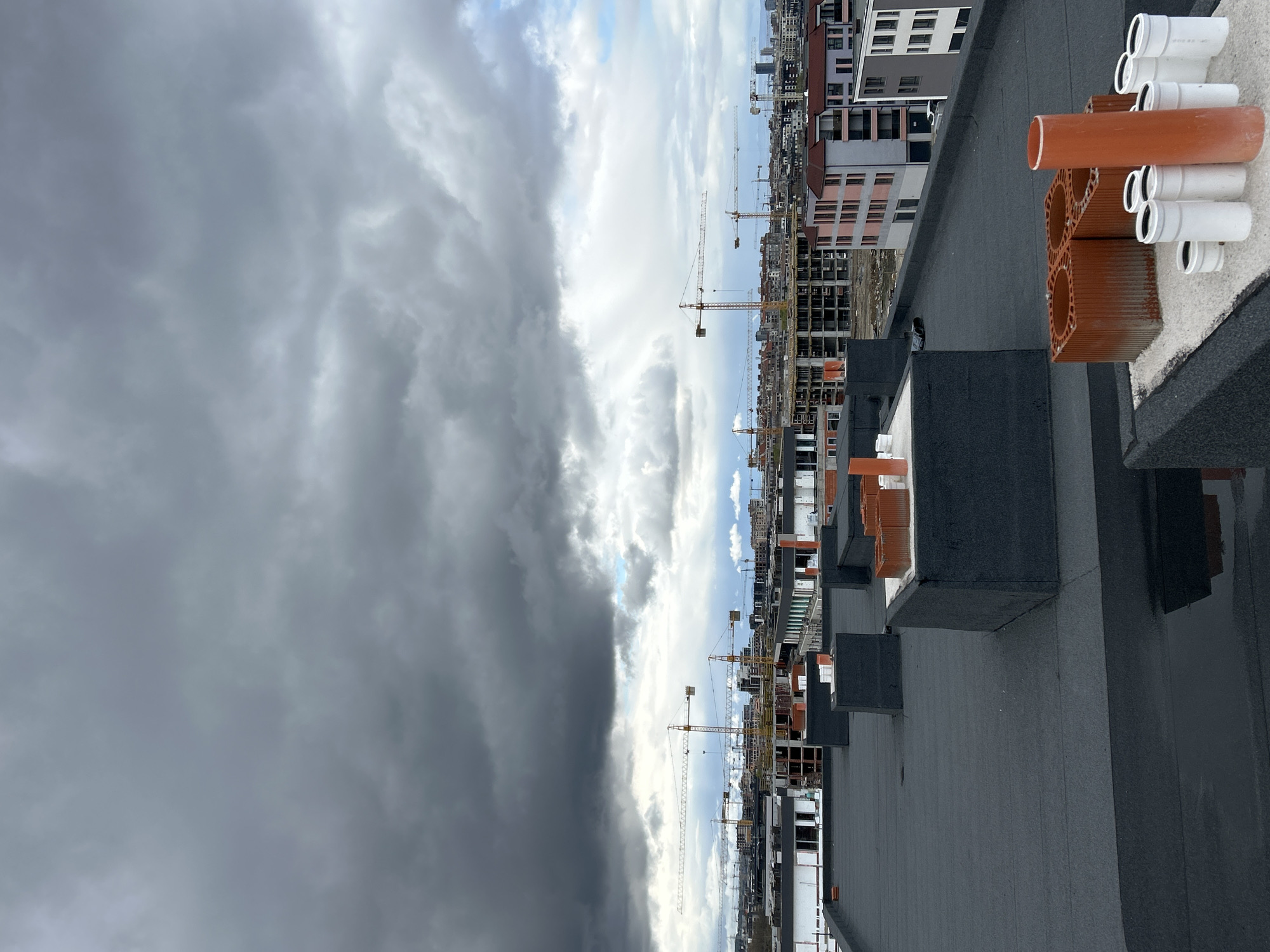 The image depicts a view from a high vantage point, possibly a rooftop, overlooking a construction site in what appears to be an urban area, likely Sofia, Bulgaria, based on the architectural style and construction activity visible. The sky is partially covered with dark, ominous storm clouds on one side and brighter, scattered clouds on the other.
In the foreground, on the rooftop, there are several sections of orange and white PVC pipes and other construction materials. The rooftop itself is covered with dark gray bitumen membrane.
In the middle ground, a series of buildings are under construction. Several construction cranes are visible amongst the buildings, indicative of ongoing development. The architecture of the partially constructed buildings suggests a modern design with a mix of gray and white facades.
In the background, the cityscape extends into the distance, with more buildings visible. The overall scene suggests a period of dynamic construction activity juxtaposed against a potentially stormy weather condition. There are no people visible in the image.