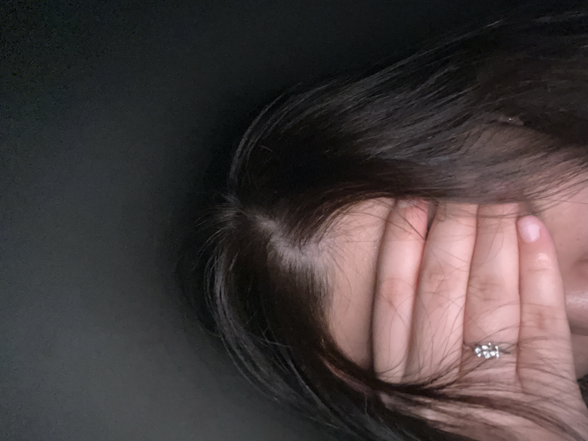 A close-up, eye-level photograph features an individual covering their upper face with their hand in the city of Unknown, Unknown. The individual has dark brown hair, parted slightly off-center, revealing a light-toned scalp. A hand with light, slightly pinkish skin and short, unpainted nails is positioned horizontally across the face, concealing the eyes and upper nose. A silver-toned ring with a cluster of small, clear stones in a floral or geometric design is worn on the ring finger of the covering hand. Only the forehead above the hand and a small portion of the lower face around the hand are discernible. The background is uniformly dark and devoid of any identifiable features.
