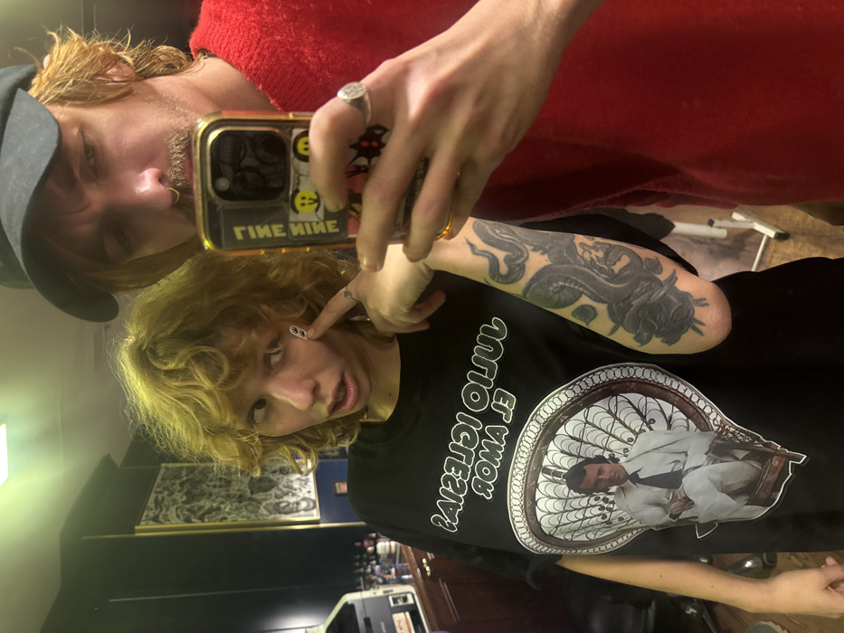A selfie captures two people. The person in the foreground has curly blonde hair, a black t-shirt with an image of a man and text, and a "33" sticker on their face. The person taking the photo is wearing a black cap and a red sweater. They hold a phone with a gold case. A tattoo is visible on the arm. The background appears to be a room with furniture and framed artwork.
