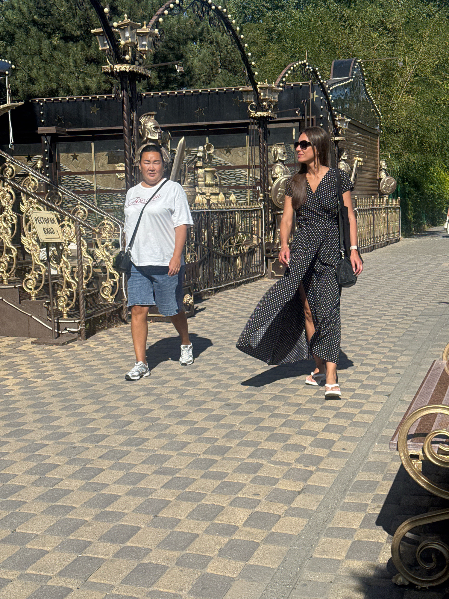 Two women are walking on a paved sidewalk. The woman on the left is wearing a white t-shirt and denim shorts, with a black crossbody bag. The woman on the right is wearing a black and white polka dot maxi dress with a high slit, sunglasses, and a black shoulder bag. They are in front of a building with a dark facade and ornate golden railings. A sign in Cyrillic on the left reads "РЕСТОРАН ВХОД", which translates to "RESTAURANT ENTRANCE". The environment appears to be a park or outdoor public space in the city of Anapa, Russia. The sidewalk is paved with a checkerboard pattern of beige and grey tiles.