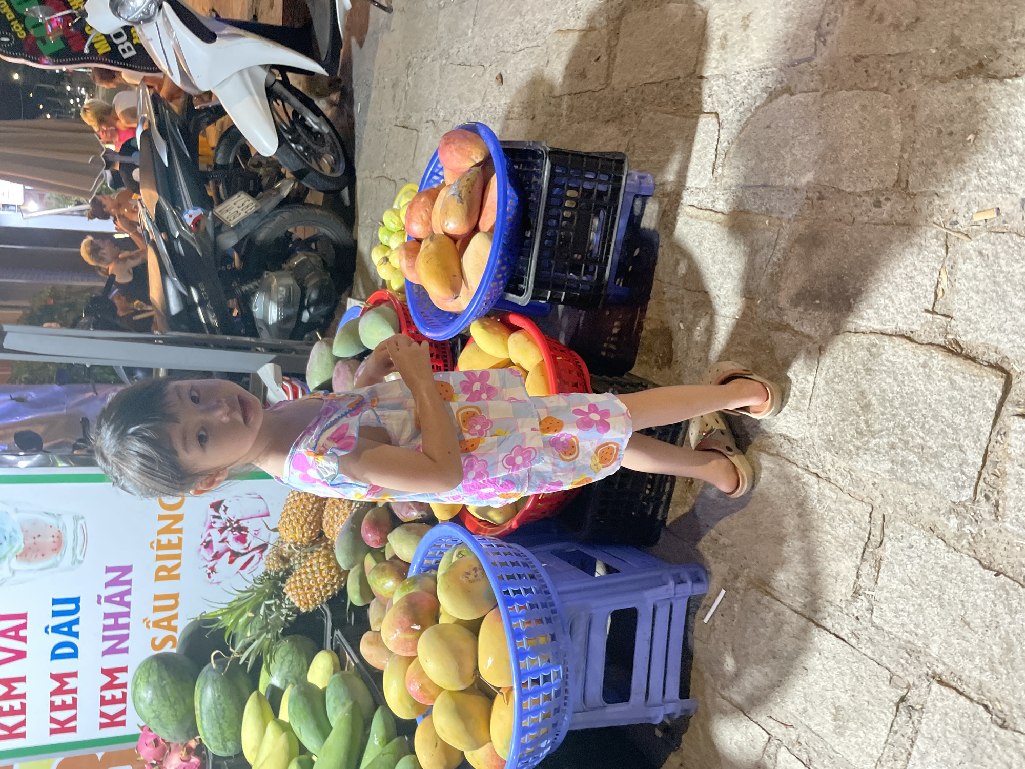 A young girl stands beside a display of fruits in Phu Thuy Ward, Vietnam. The fruits, which include mangoes, watermelons, and pineapples, are piled in blue and red plastic baskets. Behind the display, a sign advertises "Kem Vai, Kem Dau, Kem Nhan, Sau Rieng," indicating that the vendor sells ice cream as well. Parked motorcycles are in the background, and the ground is paved with uneven stones.