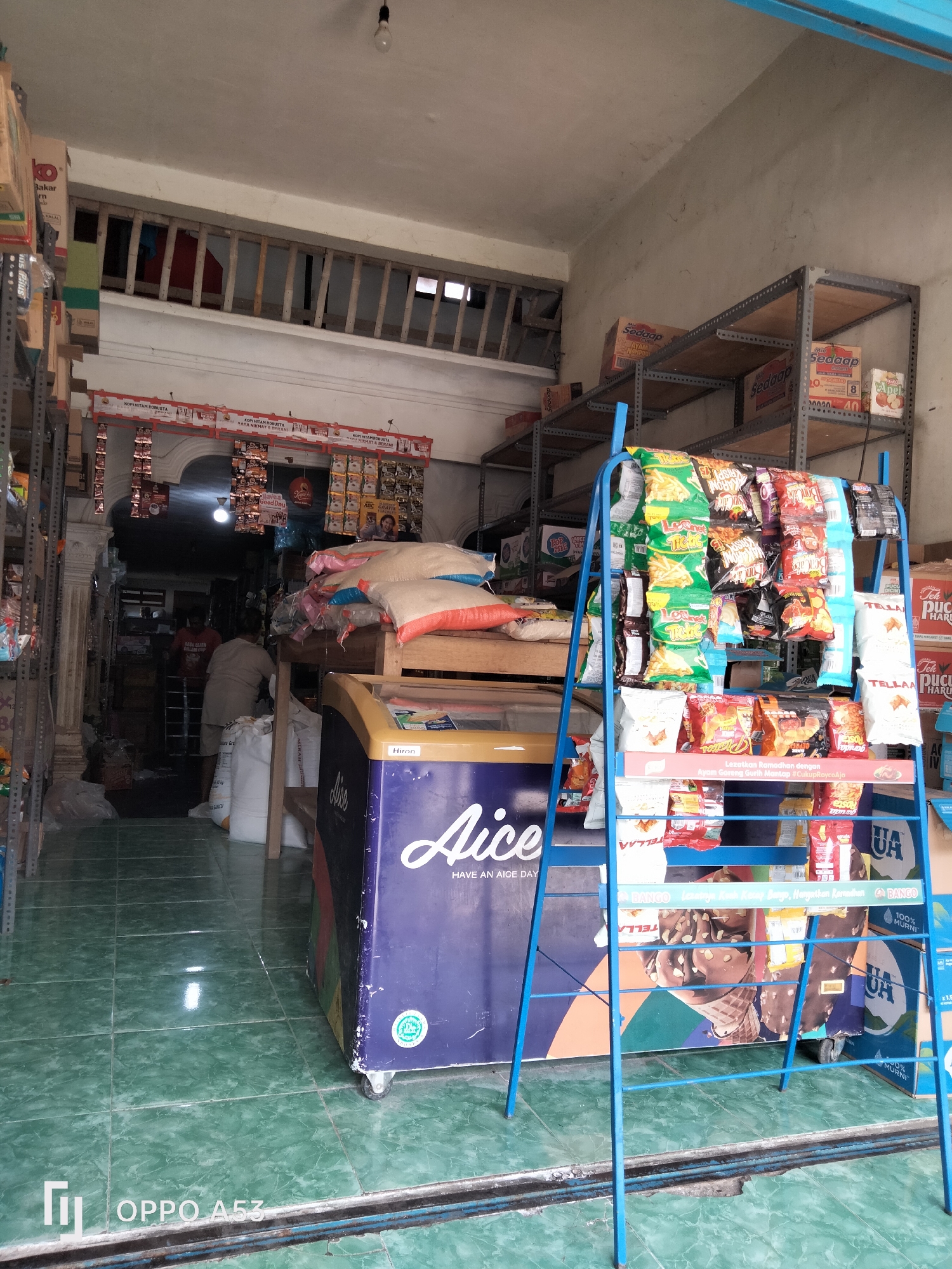 The image depicts the interior of a small convenience store located in Kelutan, Indonesia. The store appears to be somewhat cluttered with merchandise organized on shelves and display racks. In the foreground, a blue metal rack is filled with various bagged snacks, some of which are brightly colored. To the right of the snack rack, a blue and purple branded freezer is visible, with the brand name "Aice" clearly displayed. Beyond the freezer, large sacks of rice are stacked. Further into the store, a person wearing a light-colored shirt and shorts is seen interacting with merchandise. Another person in a red shirt is partially visible behind them. The floor is tiled in a green pattern, and the walls are a neutral, textured color. Shelving units are present throughout the store, holding a variety of packaged goods, including boxes of instant noodles and other food items. The overall impression is of a functional, albeit modestly equipped, local retail establishment.