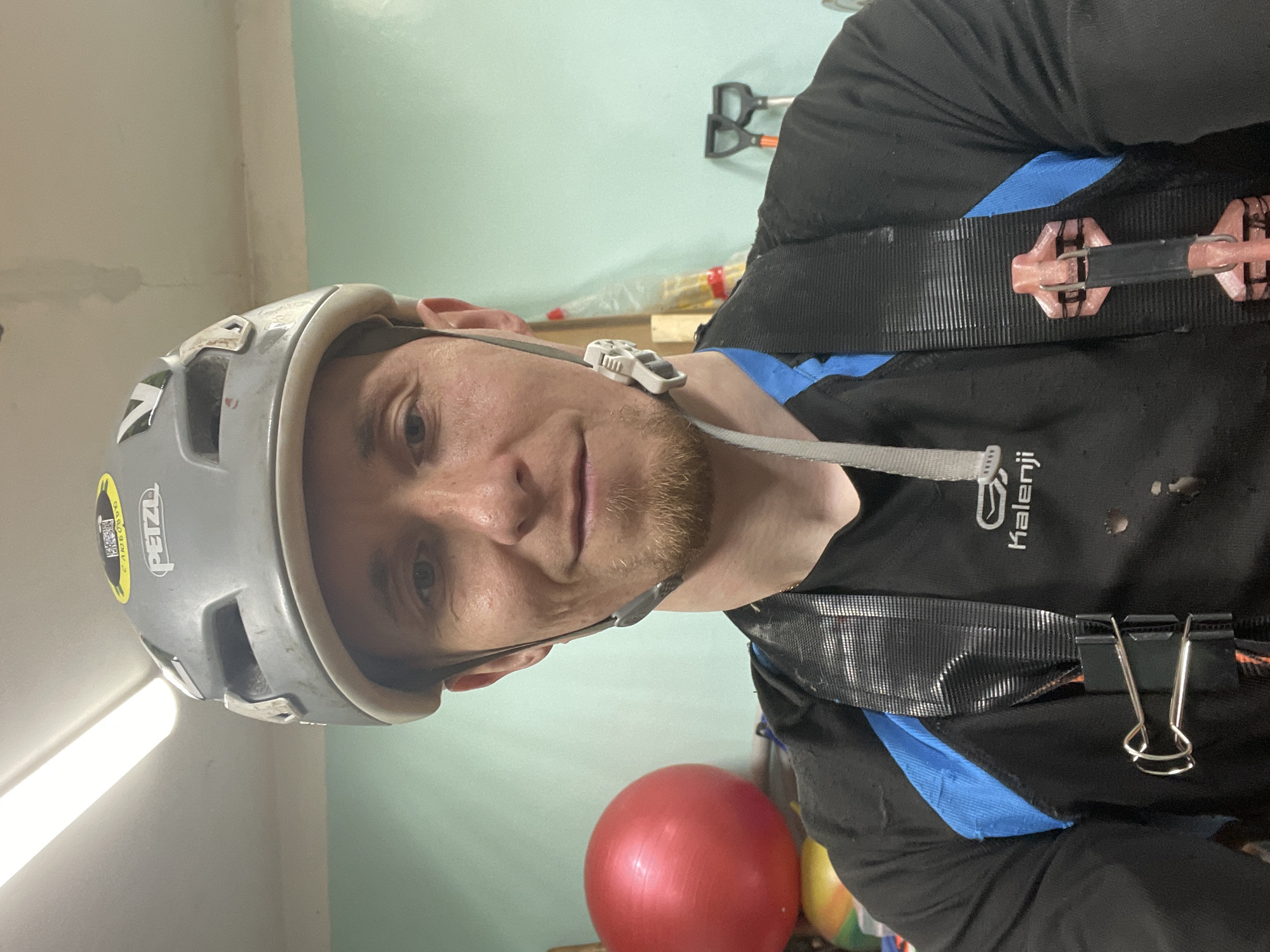 A man wearing a grey helmet with a Petzl sticker and a climbing harness poses for a selfie indoors. He is wearing a black and blue shirt. There's a red exercise ball visible in the lower part of the frame and a shovel mounted on the wall in the background. The location is unspecified, and no mention of Sarapul, Russia can be found in the image.