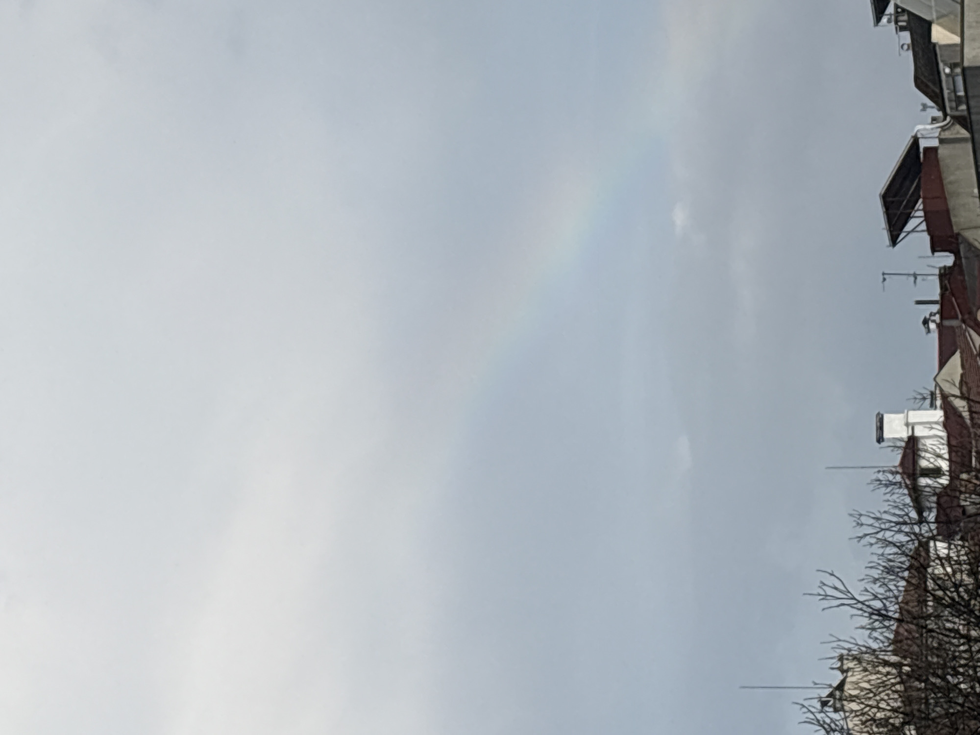 The image shows a cloudy sky with a faint rainbow arching across it. In the bottom right corner, the tops of buildings and bare tree branches are visible. Based on the architectural style and the presence of bare tree branches (suggesting a colder season), the location might be the city of Burgas, Bulgaria, given the user's prompt. However, without additional cues, the specific location within Burgas remains unclear. The general scene suggests a neutral, overcast day.