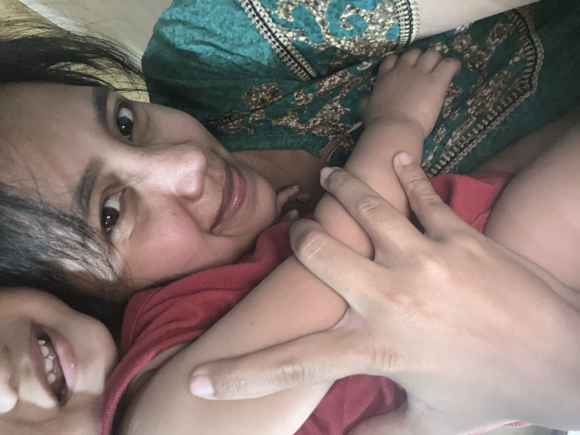 This media file captures a close-up, overhead shot of a woman and two children, likely a mother and her children, in an intimate embrace. The subjects are positioned close together, with the woman's face in the upper portion of the frame and the children nestled against her. The woman has dark hair and is looking directly at the camera with a gentle smile. She is wearing a green garment with a patterned print. One child, wearing a red garment, is partially visible in the lower left of the frame, their mouth open in a smile. Another child's hand is visible on the woman's arm, suggesting they are also being held. The overall mood is one of warmth and affection. The setting appears to be indoors, possibly a bedroom or living area, given the soft lighting and the presence of fabric. The time of day is not explicitly indicated, but the lighting suggests it could be daytime. The location context of Depok, Indonesia, provides a cultural backdrop that might inform the attire and overall aesthetic, though no specific landmarks or explicit cues are visible. There is no visible text in the image.