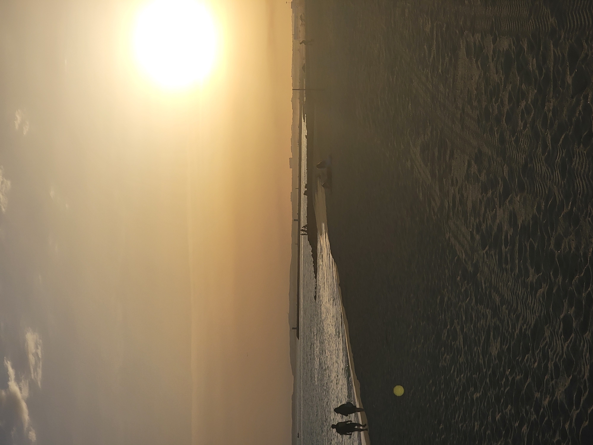 The image shows a wide, sandy beach at sunset in Quarteira, Portugal. The sun is low in the sky, casting a warm, golden glow over the scene. The sky is a hazy orange and yellow, with a few wispy clouds. The ocean is calm, with gentle waves lapping at the shore.
In the foreground, two people are walking along the waterline, their silhouettes dark against the bright sand. Further down the beach, other figures can be seen, some sitting, some standing, their forms indistinct in the dim light. A bright yellow object, possibly a ball, is visible on the sand in the lower left quadrant of the image.
In the distance, a long, breakwater-like structure juts out into the sea, with a lighthouse visible at its end. The coastline stretches out, with buildings and hills forming a faint outline against the horizon. The overall impression is one of tranquility and a peaceful end to the day.