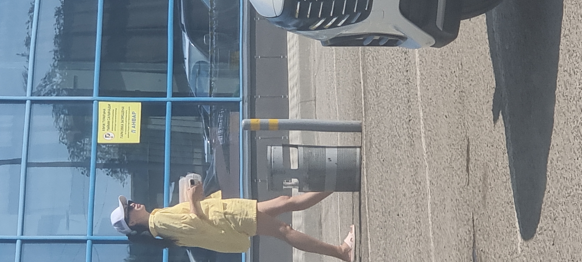 A young woman wearing a yellow shirt, yellow shorts, a white baseball cap, and pink sandals walks from left to right in front of a modern building with blue-tinted windows in Aktobe, Kazakhstan. She is holding a light-colored container in her right hand. A yellow sign is attached to the building's windows, displaying text in Cyrillic and Latin alphabets, with a parking symbol. A cylindrical trash receptacle with horizontal bands is located to the woman's right. A grey pole with yellow bands stands to the right of the trash receptacle. The ground is paved with asphalt and marked with white lines. The front right corner of a silver vehicle is visible in the lower right portion of the image.