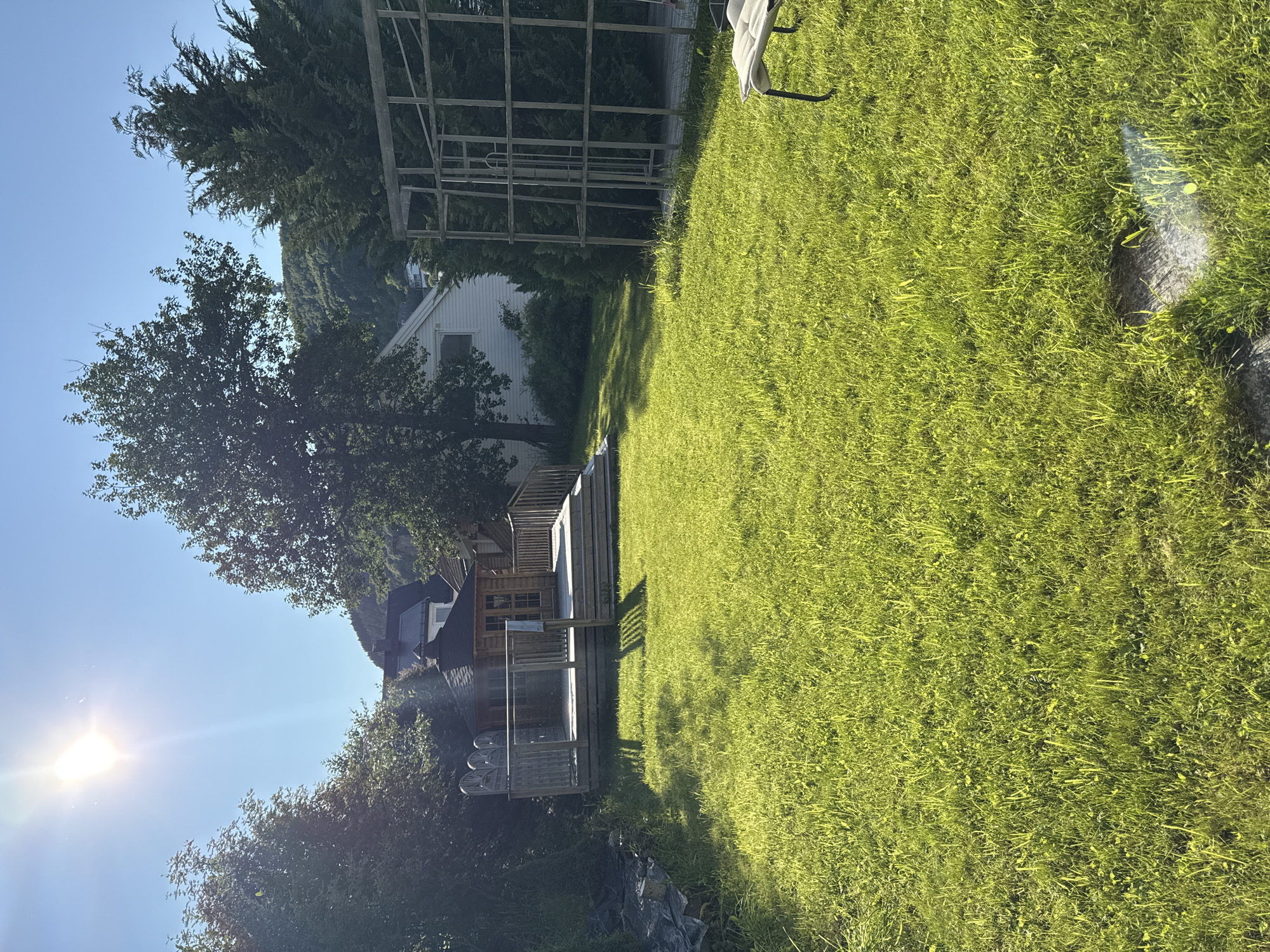 The image depicts a sun-drenched backyard with lush green grass. In the background, a wooden deck with a small cabin-like structure is visible, possibly a gazebo or playhouse. Beyond the deck, a white house with dark roofing stands, and further up a hill, trees and a distant structure are seen against a clear blue sky. To the right of the yard, a metal frame, potentially a garden trellis or part of an unfinished structure, is present. A white chair with black legs is positioned on the grass in the far right of the image. The sun is shining brightly, casting long shadows across the lawn. The overall scene suggests a residential setting in Harstad, Norway, likely during a sunny day.