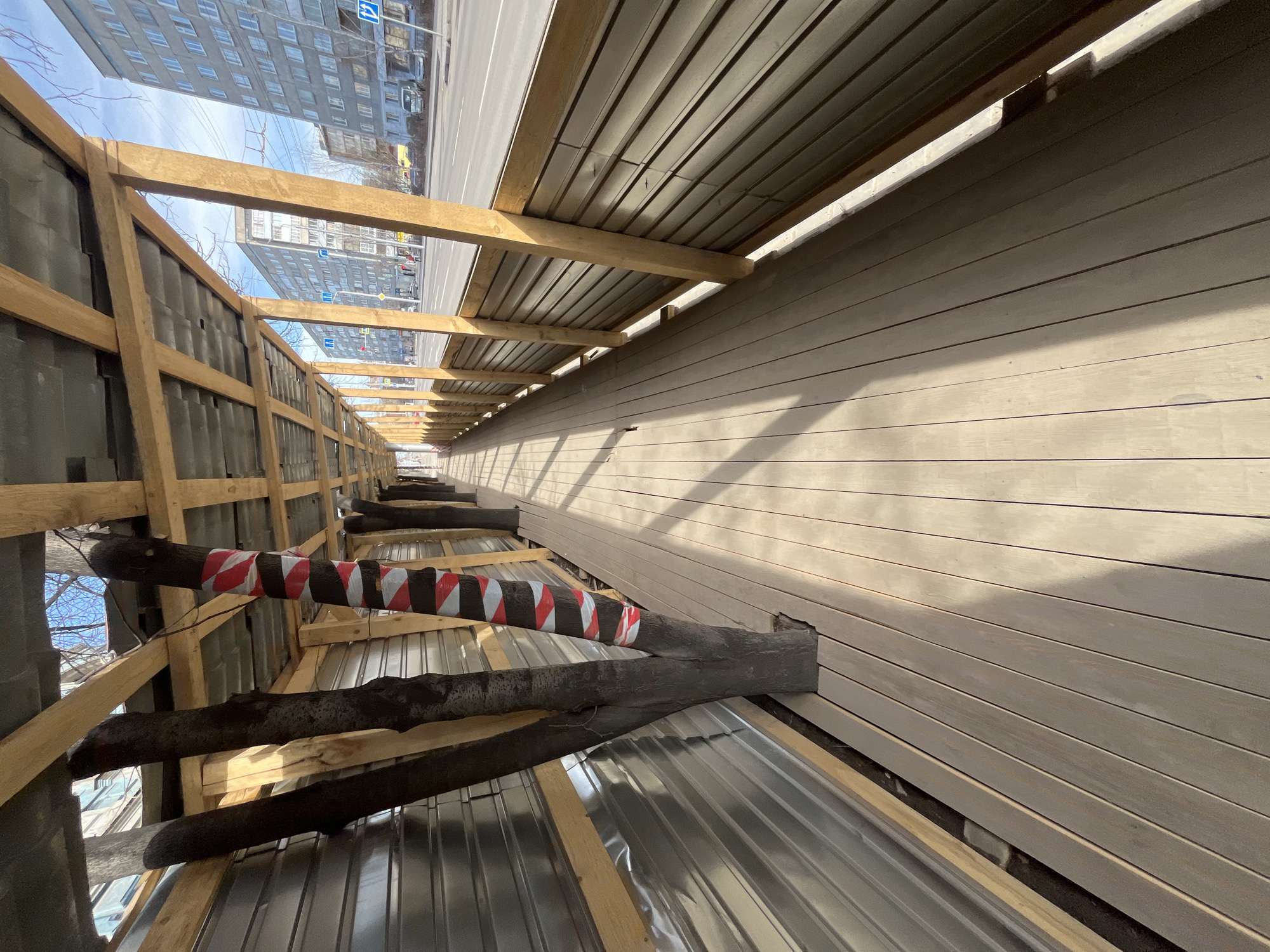 The image shows a construction site in Unknown, Unknown, featuring a partially erected pedestrian tunnel adjacent to a city street with high-rise buildings in the background. The tunnel structure consists of a wooden frame with metal sheeting as a roof and wall paneling on one side. Tree trunks, some wrapped in red and white striped tape, act as supports for the structure. The street visible beyond the tunnel frame indicates a clear sky above the urban environment.