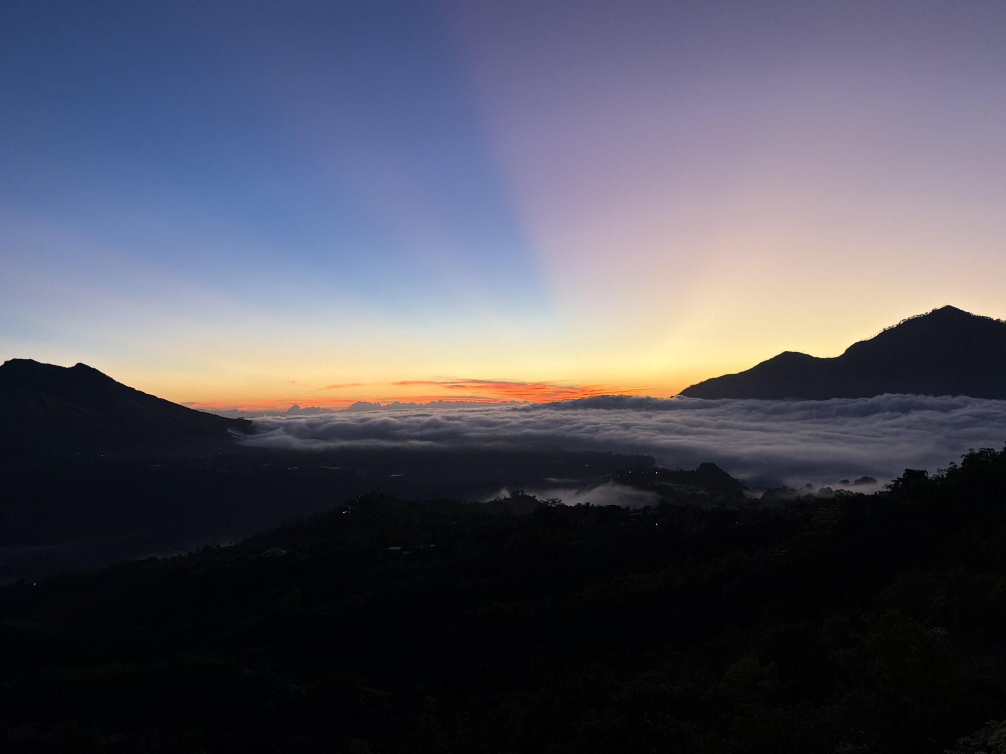 The image depicts a serene pre-dawn landscape viewed from a vantage point in Kintamani, Indonesia. Silhouetted mountains frame the scene, with the left side featuring a dark, rugged peak against a gradient sky of deep blue transitioning to soft purple and then a vibrant band of orange and yellow near the horizon. Streaks of light, resembling crepuscular rays, emanate upwards from the horizon, piercing the twilight sky.
Below the horizon, a thick layer of clouds or mist blankets the valley, obscuring the terrain and creating an ethereal, sea-of-clouds effect. Several indistinct, dark shapes of hills or smaller mountains emerge from this cloud cover. Scattered lights, likely from a settlement, are visible within the lower, darker foreground and also subtly within the misty valley, indicating the presence of human habitation. The foreground consists of dark, silhouetted slopes covered in vegetation, adding depth to the composition. There are no visible people or specific actions taking place in the scene; it is a static, environmental capture. The overall atmosphere is one of tranquility and the quiet anticipation of sunrise.