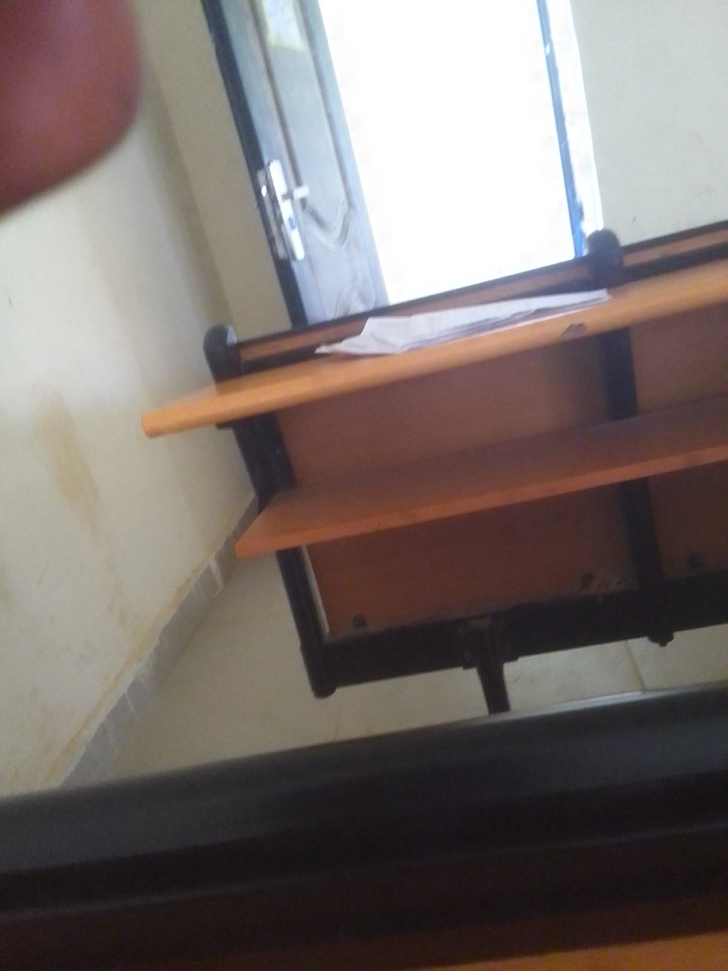 The image shows a close-up view of a wooden desk with black metal supports, likely in a classroom or office setting. The desk has at least two shelves, both made of a light brown wood. There are some papers placed on the top shelf. The background shows a wall painted a light cream color with some yellow stains and a doorway with a metal handle. The lighting suggests it is daytime, possibly indoors with natural light coming from a window or open doorway, creating a bright glare in the upper right portion of the image. The perspective is from a low angle, looking upwards at the desk. No people are visible. The overall impression is of an ordinary, functional workspace.