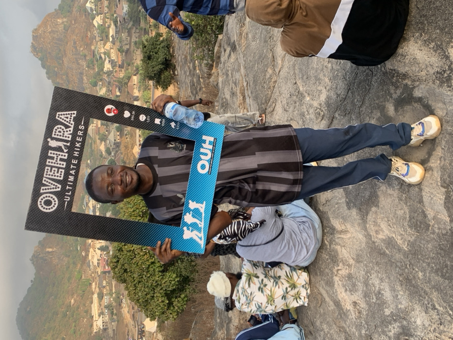 A group of individuals is positioned outdoors on a rocky elevation overlooking the city of Okengwe, Nigeria, nestled amidst rocky hills. A male individual stands centrally in the foreground, facing the camera with a slight smile. He is dressed in a grey and black striped short-sleeved shirt, dark blue athletic trousers with white side stripes, and light-colored sneakers. He holds a large black and blue promotional frame, inscribed with "OVEHIRA - ULTIMATE HIKERS -" at the top and "OUH" with hiker silhouettes at the bottom, along with social media engagement metrics (heart, comment, share, save, profile, 245.2K, 1,277, 8,939). A clear plastic water bottle is held in his right hand. Multiple other individuals are present; some are seated on the grey, weathered rock surface, including one wearing a white beanie and a light-colored, palm-leaf patterned shirt, and another clad in a brown, white, and black color-block hooded garment. The background reveals a residential settlement within Okengwe, Nigeria, characterized by numerous buildings with light-colored roofs, situated at the base of and extending up several verdant, rocky hills, all under a brightly lit sky.