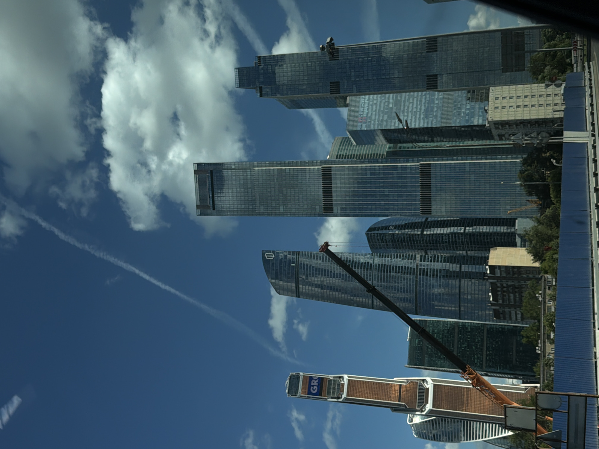 This media file captures a view of modern skyscrapers against a bright blue sky with scattered white clouds. The buildings are tall and feature glass facades, reflecting the sky and surrounding environment. In the foreground, a construction crane extends upwards, with its boom partially obscuring one of the buildings. The crane's arm is dark, and its hook is visible at the top. Below the crane, a structure with the word "GRO" visible in blue letters on a white background is also present, likely part of a construction vehicle or equipment. The overall scene suggests an urban setting, likely Moscow, Russia, given the architectural style and common presence of construction in such a metropolis. The lighting indicates it is daytime, possibly late morning or early afternoon, with clear weather conditions. The perspective is from a slightly elevated viewpoint, looking upwards at the towering structures.