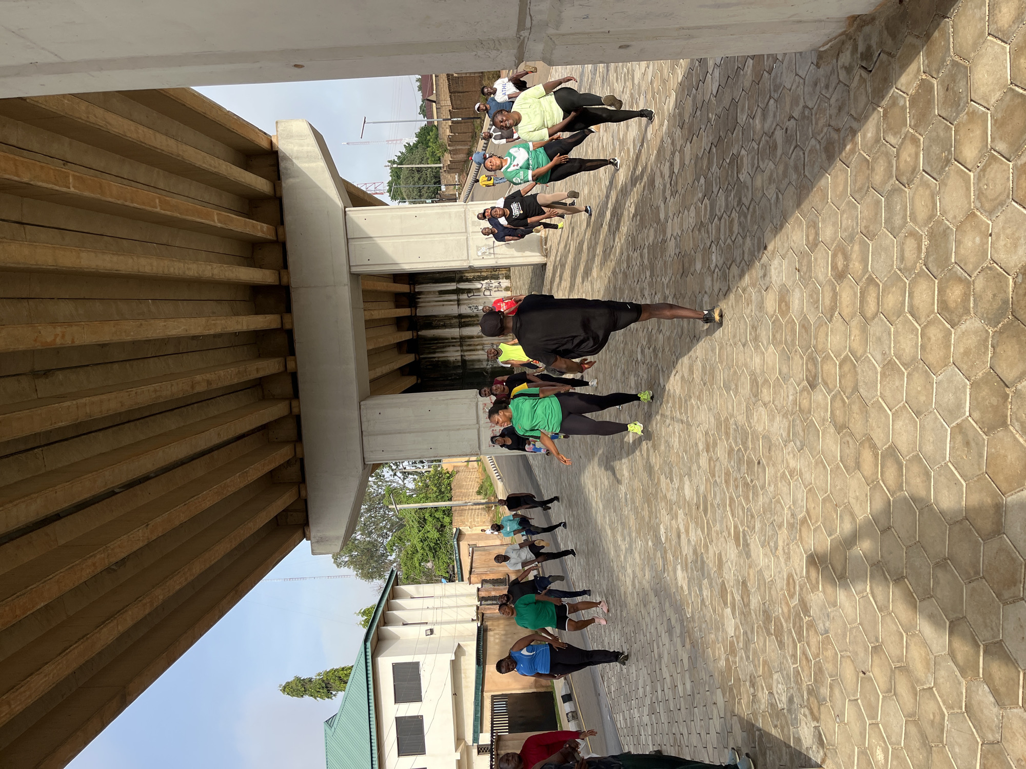 The image depicts a group of approximately twenty individuals engaged in an outdoor exercise routine beneath an elevated concrete highway structure. The scene appears to be in Jos, Nigeria, given the architectural style of the nearby buildings, the vegetation, and the presence of a distinct communication tower in the background.
The ground is paved with interlocking hexagonal concrete blocks, casting long shadows from the overhead structure and the participants. The participants, varying in age and gender, are dressed in athletic wear, including t-shirts, leggings, shorts, and athletic shoes. They are performing coordinated movements, suggestive of a warm-up or stretching exercise, with many lifting their knees towards their chests.
In the middle ground, a white, multi-story building with a green roof is visible, situated to the left of the highway structure. Adjacent to it are fences and more vegetation. The sky is a clear, pale blue, indicating daytime, likely morning or late afternoon due to the angle of the shadows. The overall atmosphere is one of communal physical activity in an urban, infrastructural setting.