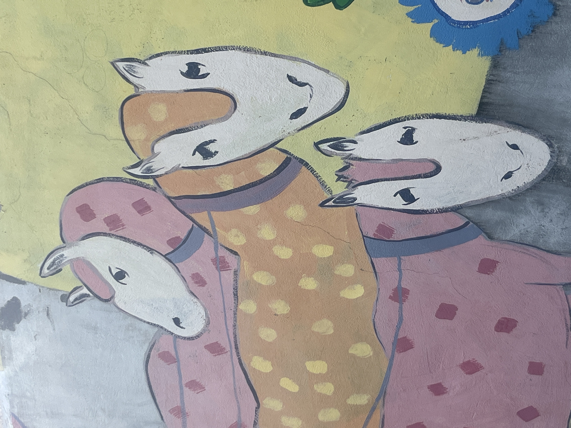 The image shows a section of a colorful mural on a weathered wall, likely outdoors. The mural depicts three stylized horses with simplified features, painted in shades of pink, orange, and white, with dark outlines. The horses have distinct patterns: one has pink polka dots, another has yellow polka dots, and the third has pink square accents. A partially visible blue flower with a white center is also present on the right side of the mural. The background of the mural is a patchy mix of yellow and grey, and the wall itself appears to be aged, with visible cracks and texture. The artwork seems to be located in an urban environment, given the nature of the wall and the mural. Without further information or context, the specific location in Kyiv, Ukraine cannot be determined.