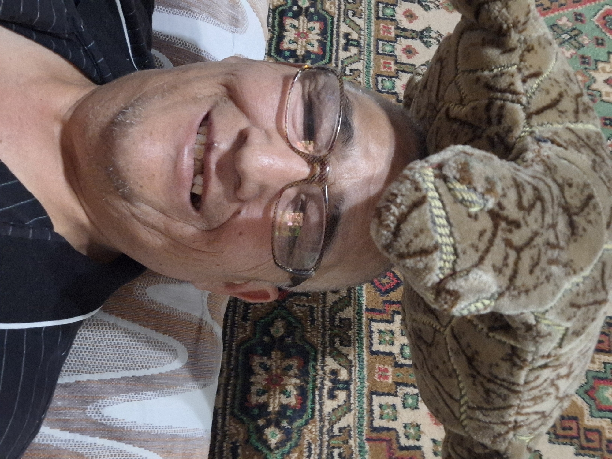 An individual, wearing glasses and a black and white striped shirt, is photographed from the chest up, with a plush toy positioned on top of their head. The background consists of a patterned rug and a textured cushion. The individual's facial expression appears to be a smile, revealing missing teeth. The image is a selfie, taken indoors. There is no information available about the specific location within Almaty, Kazakhstan.