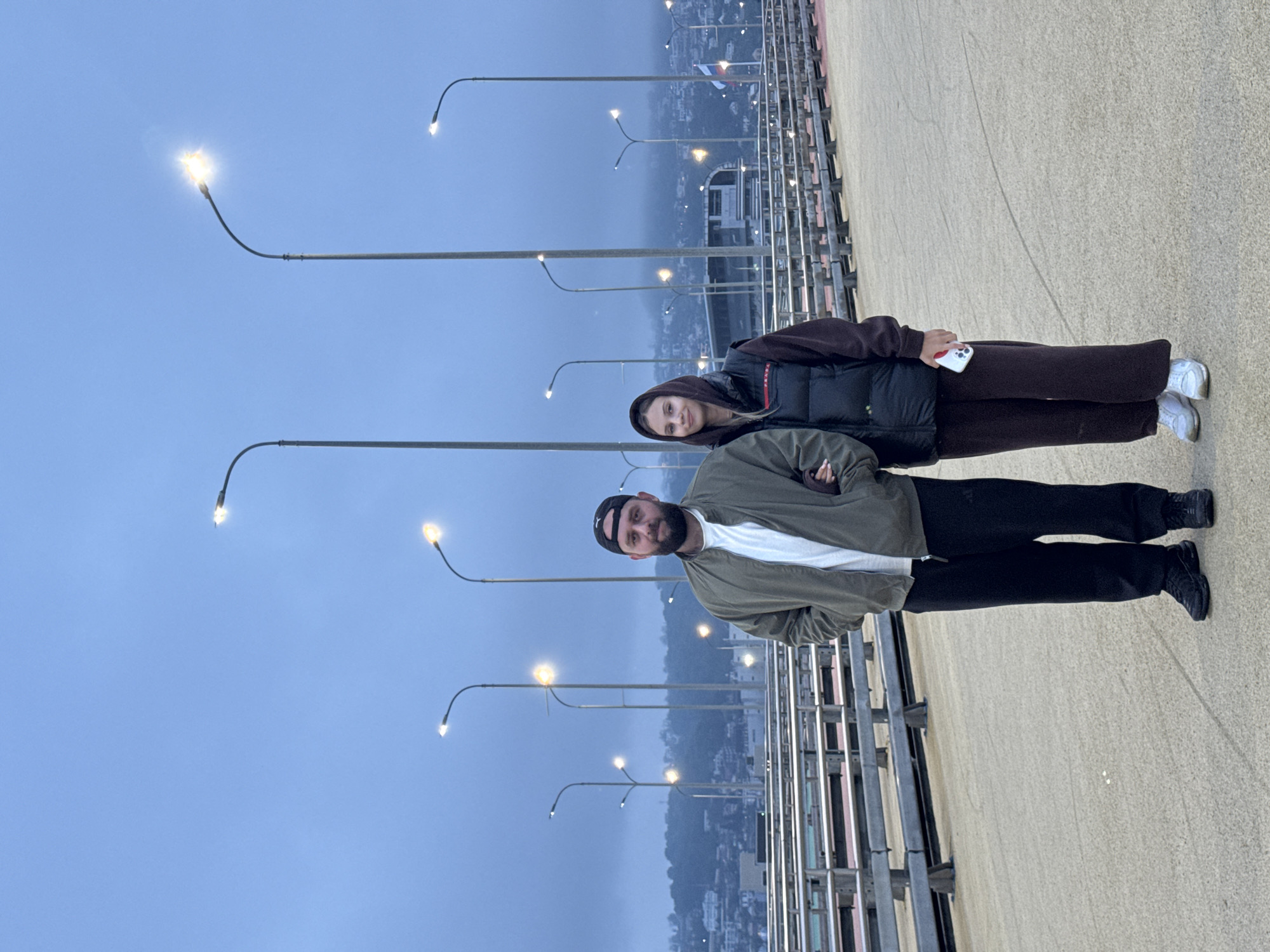 The image, correctly oriented, depicts a man and a woman posing on what appears to be a bridge or elevated walkway at dusk or early night. The sky is a deep, hazy blue, and numerous streetlights with bright, starburst-like glows illuminate the scene, extending into the distant background.
On the right side of the frame, a man stands, facing slightly towards the camera with a faint smile. He has a beard and is wearing a black beanie, a white t-shirt, an olive-green jacket with crossed arms, black pants, and dark shoes. Behind him and to his left, a woman is positioned, also smiling towards the camera. She has blonde hair and is dressed in a dark brown hooded jacket over a black puffer vest, dark brown wide-leg pants, and white sneakers. She holds a white smartphone with a red case, displaying a visible Apple logo, in her right hand.
To the left of the subjects, a metal railing system lines the edge of the walkway. Beyond the array of streetlights, a sprawling city or town is visible in the mist, with hills or mountains in the far distance. A flag, appearing to be the Russian flag with white, blue, and red horizontal stripes, is partially visible in the background, offering a specific location cue for Sirius, Russia. Both individuals appear relaxed and content, suggesting a casual photo session in an urban setting during twilight hours.
