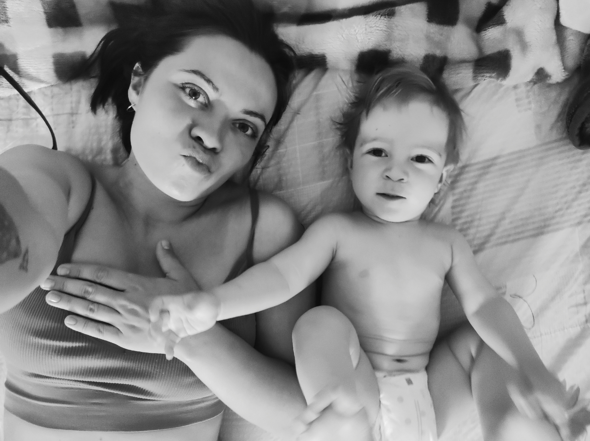 A black and white photograph depicts a woman and a child lying on a patterned surface, possibly a bed. The woman is positioned to the left of the frame, wearing a tank top, with her hand placed on her chest. Her facial expression suggests a playful pout. The child lies to the right, wearing what appears to be a diaper. Both individuals are looking directly towards the camera. The location appears to be an indoor setting.