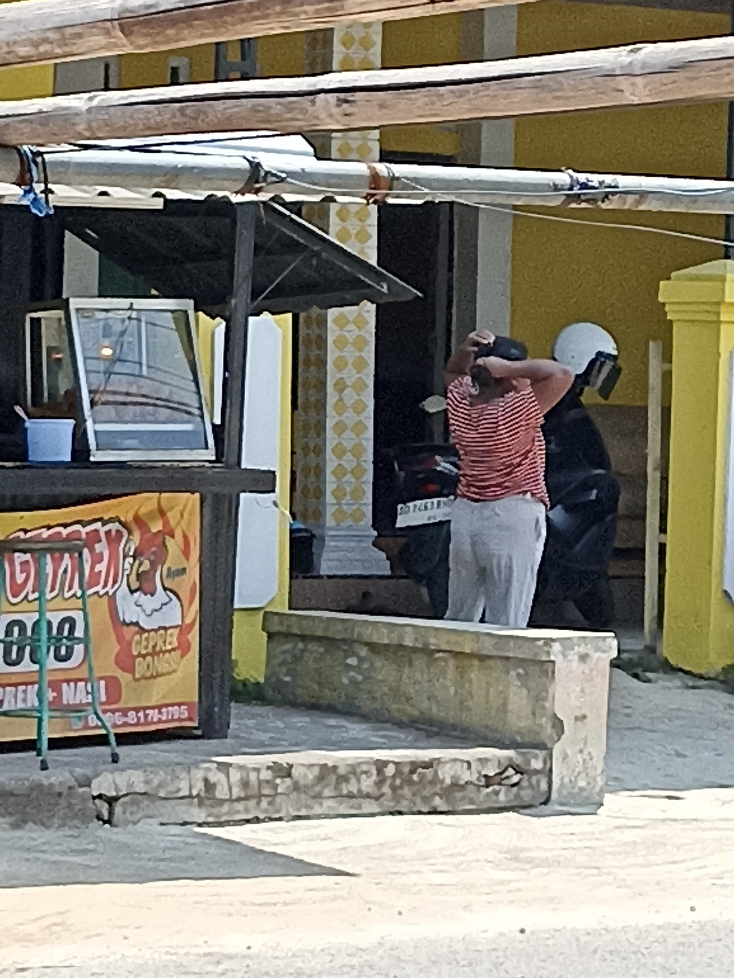 In Pulisen, Indonesia, a person wearing a red and white striped shirt and light-colored pants is standing outdoors, holding their hands up to their head as if in distress or adjusting their hair. Beside them, a person in black is wearing a white helmet. A motorcycle is parked behind them. In the foreground, a yellow sign with red lettering features a cartoon chicken and text that appears to advertise food. The scene is set against a backdrop of yellow buildings and a street.