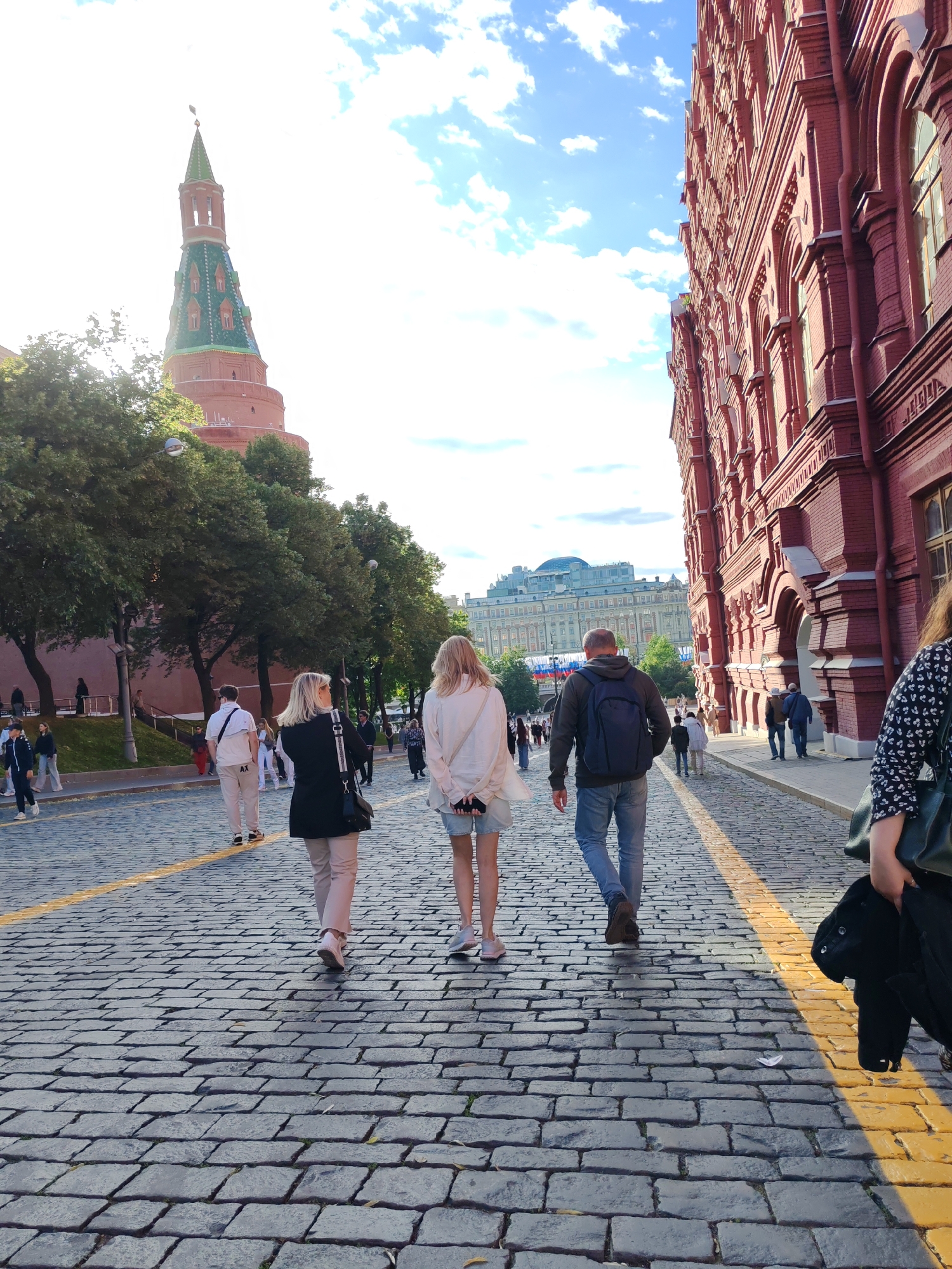 A medium shot captures a cobblestone street in an urban setting. The sun casts a warm glow, illuminating the scene from the right. In the foreground, three individuals walk away from the camera: a woman in a black jacket and light-colored pants, a woman in a white top and denim shorts, and a man in a dark hoodie and jeans with a backpack. Their backs are to the viewer as they traverse the paved street.
To the left of the frame, a distinctive red brick tower with a green, spire-like roof rises against a bright sky dotted with white clouds. Trees line the street, partially obscuring the lower portion of the tower and a reddish wall.
On the right side of the image, a tall, red brick building with numerous arched windows dominates the view. Its ornate architectural style suggests a historical or significant structure. In the middle ground, more people can be seen walking, some in smaller groups. Further in the distance, a large building with a rounded dome is visible, and flags are displayed on poles. The street is marked with a yellow line. The overall impression is one of a bustling, historic urban environment during daylight hours.