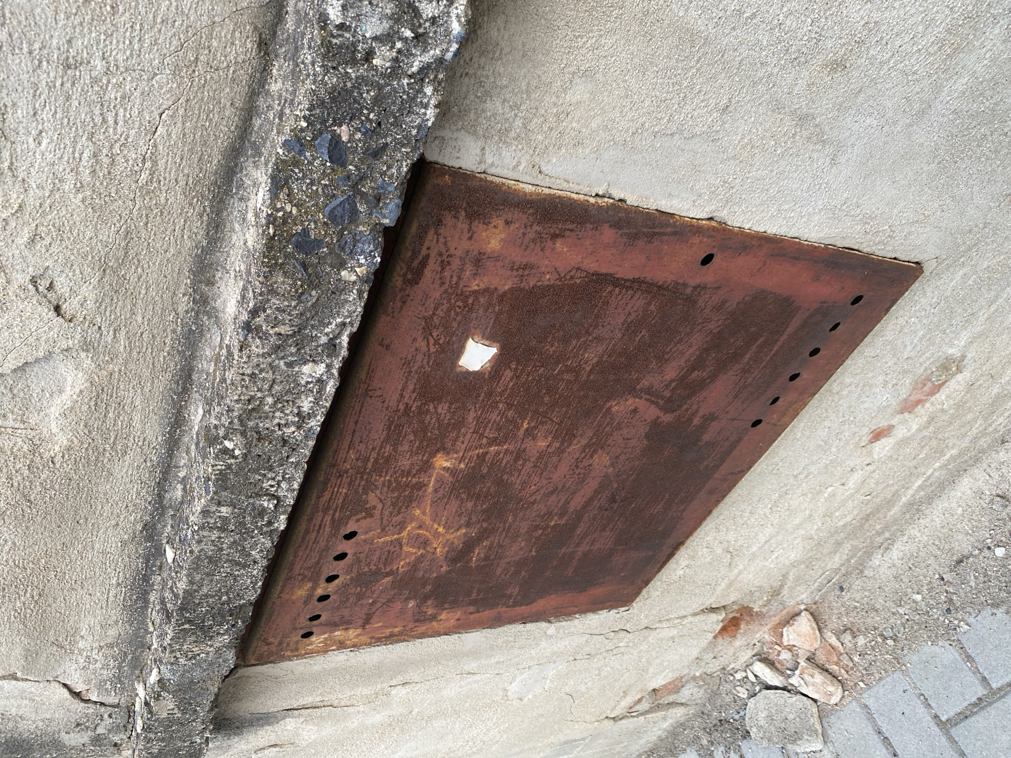The image shows a close-up of a rusted metal plate set into a concrete wall in Bílina, Czechia. The metal plate, which has several small holes along its top and left edges, appears to be a cover for some sort of utility access. The concrete wall shows signs of wear and tear, with cracks and chipped sections. A rough concrete ledge runs along the top of the metal plate. The ground in front of the wall is paved with cobblestones. The overall impression is one of age and neglect.