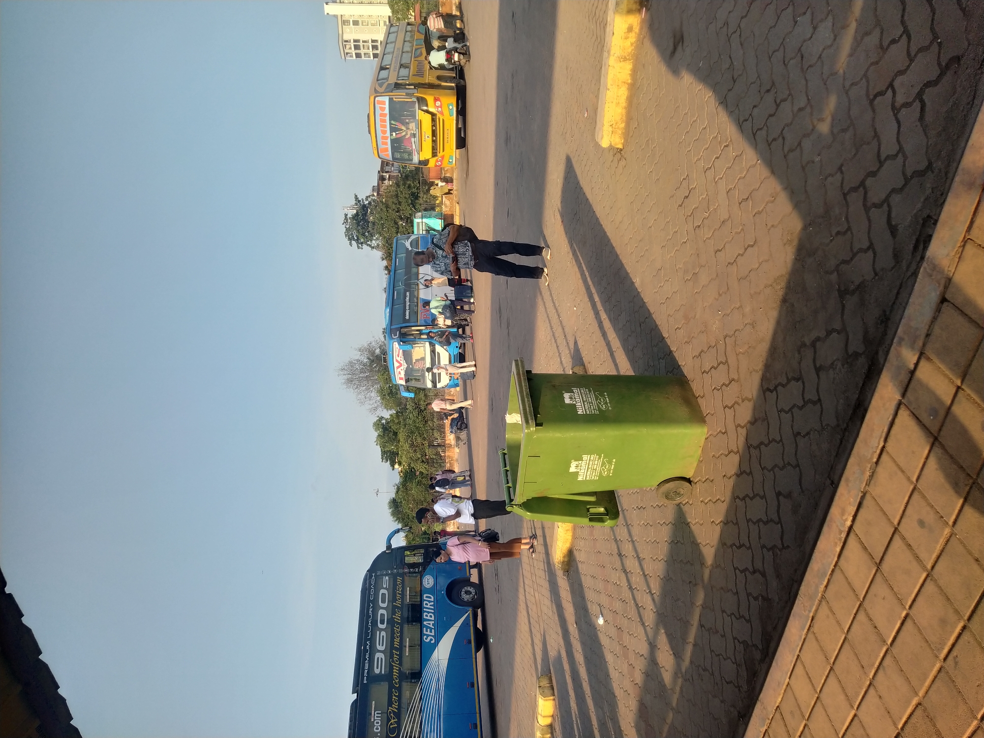 The image shows a bus station in Mapusa, India, featuring several buses lined up. A yellow double-decker bus and a blue "SEABIRD 9600s" bus are visible, along with other vehicles. People are standing and walking around the buses. A green trash bin is positioned near the edge of the paved area. The sky is clear. The ground is paved with interlocking bricks. There is a curb on the right side of the image.