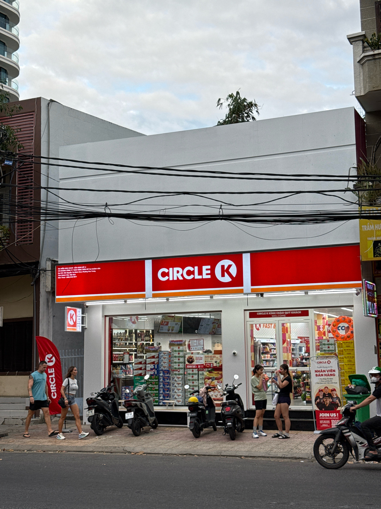 In Nha Trang, Vietnam, a Circle K convenience store is captured from the street level. The store features a prominent red and white sign with the Circle K logo. A large red banner with the Circle K logo is also visible. Inside, shelves are stocked with various products. Two young women are conversing near the entrance. Outside the store, several motorbikes are parked. Two individuals, a man and a woman, are walking on the sidewalk in front of the store. The street in front of the store is paved, and power lines are strung overhead. The sky is cloudy.