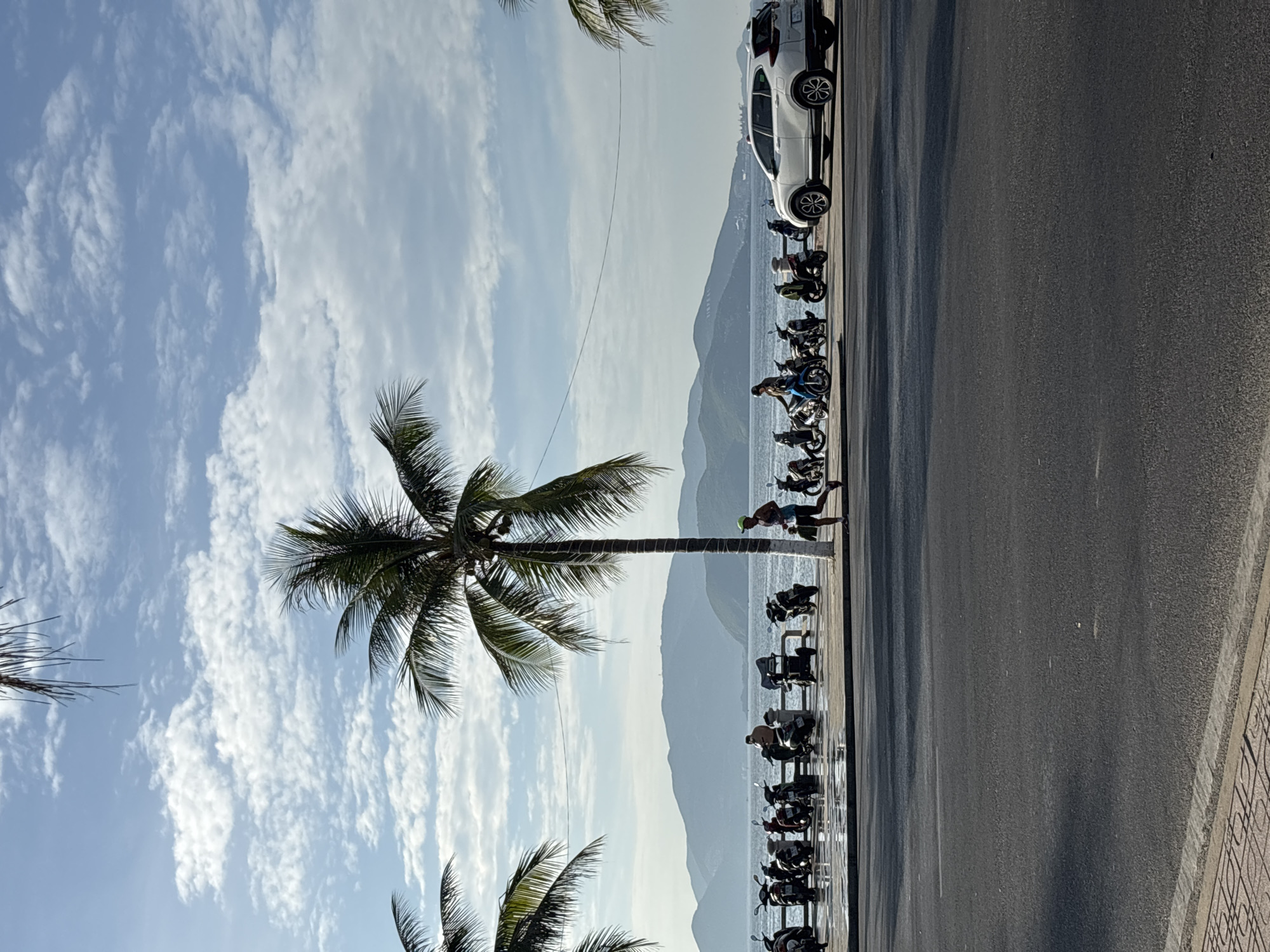 The image depicts a coastal road in Nha Trang, Vietnam. In the foreground, a paved road with a textured sidewalk is visible. Beyond the road, a walkway is lined with numerous parked motorcycles, indicating a busy area. Several palm trees are scattered along the promenade, with one prominent palm tree in the center of the frame. The sea stretches out behind the walkway, with a range of hazy mountains in the distance.
In the middle ground, a person is seen running along the walkway, wearing athletic attire and a cap. Another individual is visible on a motorcycle. A white SUV is parked to the right of the scene. The sky above is a bright blue, with scattered white clouds. The overall impression is a sunny day in a coastal city.