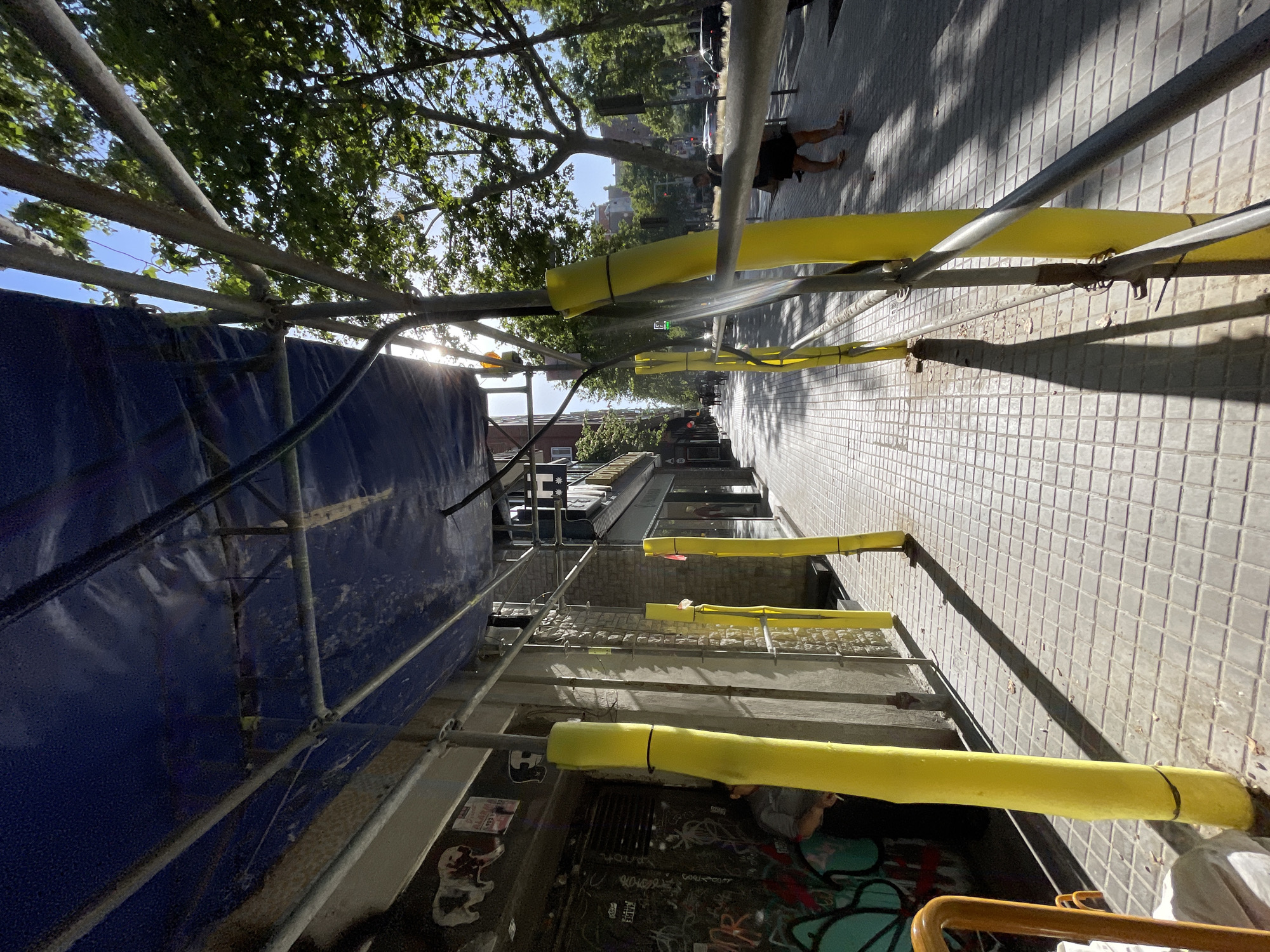 The image shows a section of a city street in Barcelona, Spain, with scaffolding and construction in progress. Metal scaffolding poles are visible, some wrapped in bright yellow padding. A large blue tarp is draped over part of the scaffolding, likely to protect the area or contain dust. The sidewalk is paved with square tiles and shows distinct shadows, suggesting it is daytime with bright sunlight. Trees line the street, their leaves filtering the sunlight and casting dappled patterns on the ground.
In the background, buildings with windows are visible, indicating an urban environment. A few people are present on the sidewalk. One person is visible from the knees down, wearing dark shorts and standing on the tiled walkway. Another person appears to be sitting or crouching near a graffiti-covered wall at the lower part of the image. The scene suggests an ongoing construction project or renovation on a city street. There is no discernible text in the image.