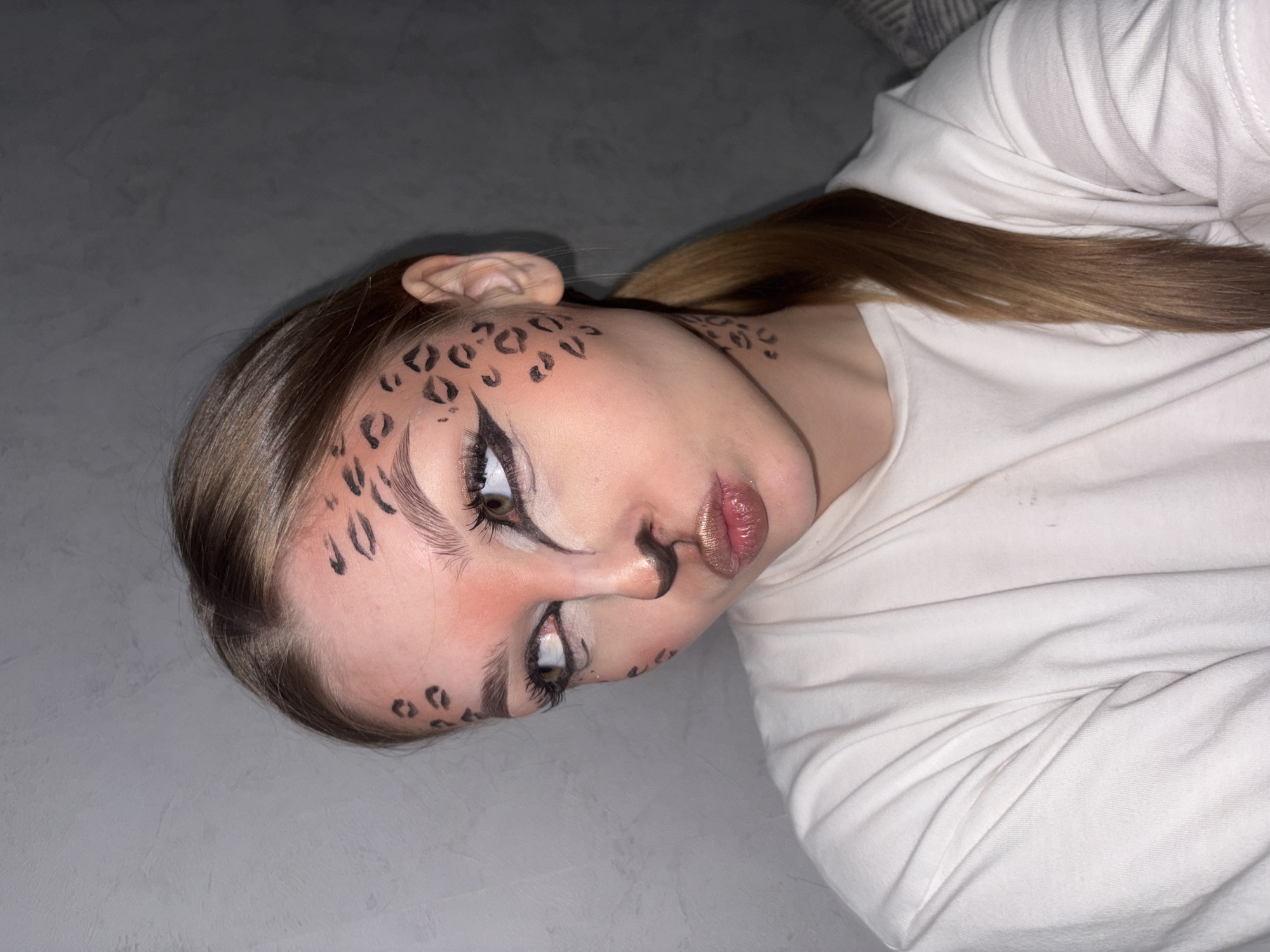 The image shows a young woman with light brown hair, styled in a low ponytail, lying on a gray surface, possibly a bed or floor. Her face is decorated with makeup, including black cheetah-like spots on her forehead and cheeks, bold eyeliner extending from the corners of her eyes, and a small black mark on her nose. Her lips are glossy. She is wearing a white long-sleeved shirt. There is no evidence indicating the location of the photo is Chaikovsky, Russia.
