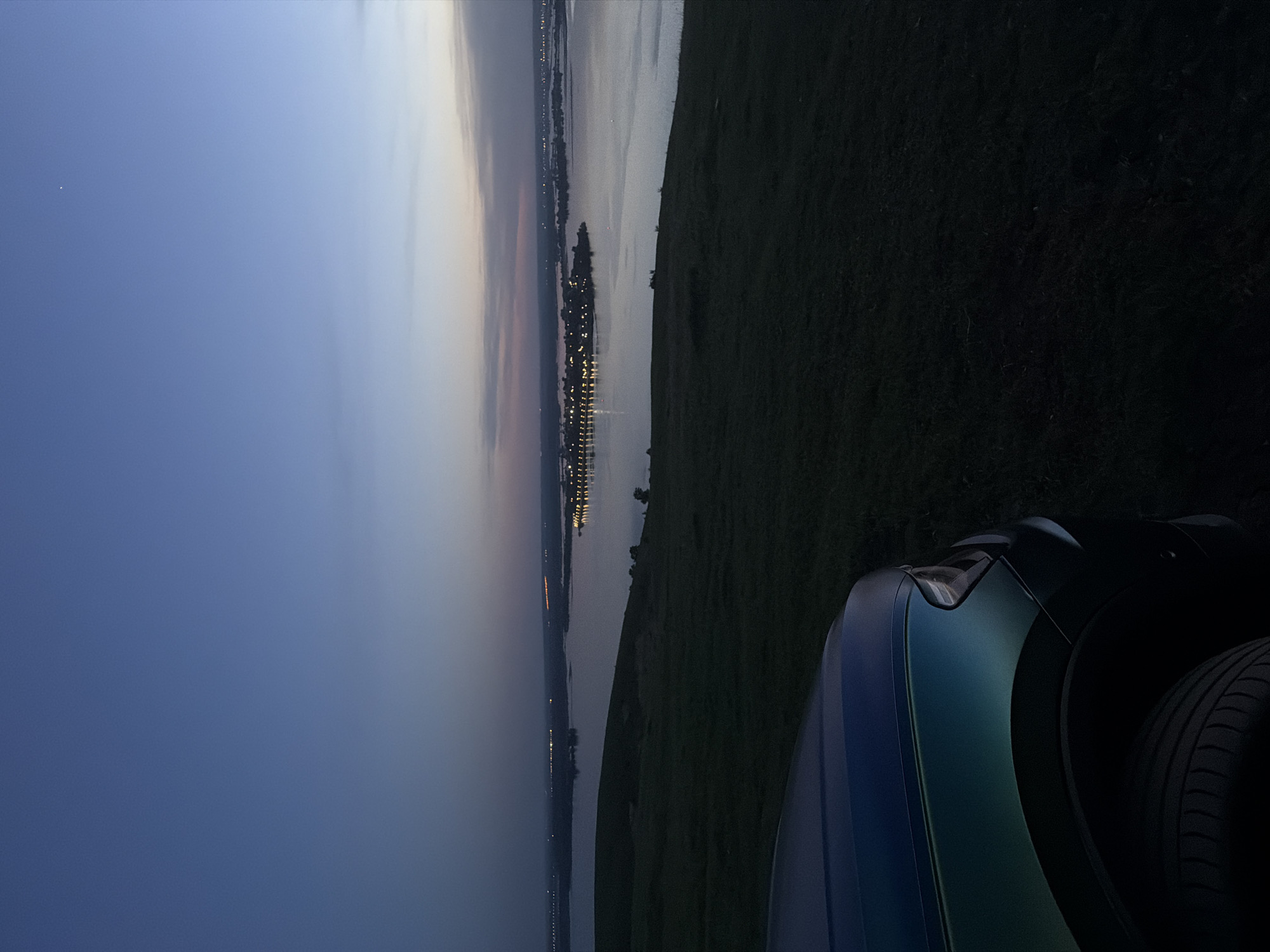 The image captures a dusky scene from Innopolis, Russia. A body of water, presumably the Volga River, stretches out with a city illuminated along its far bank. The lights suggest urban development. The sky transitions from a deep blue to a lighter hue near the horizon. A patch of grass slopes down from the right edge of the frame. The right corner of the image contains the hood and front tire of a dark-colored car. The overall setting indicates a viewpoint overlooking the city and river, possibly from a higher elevation point within Innopolis.