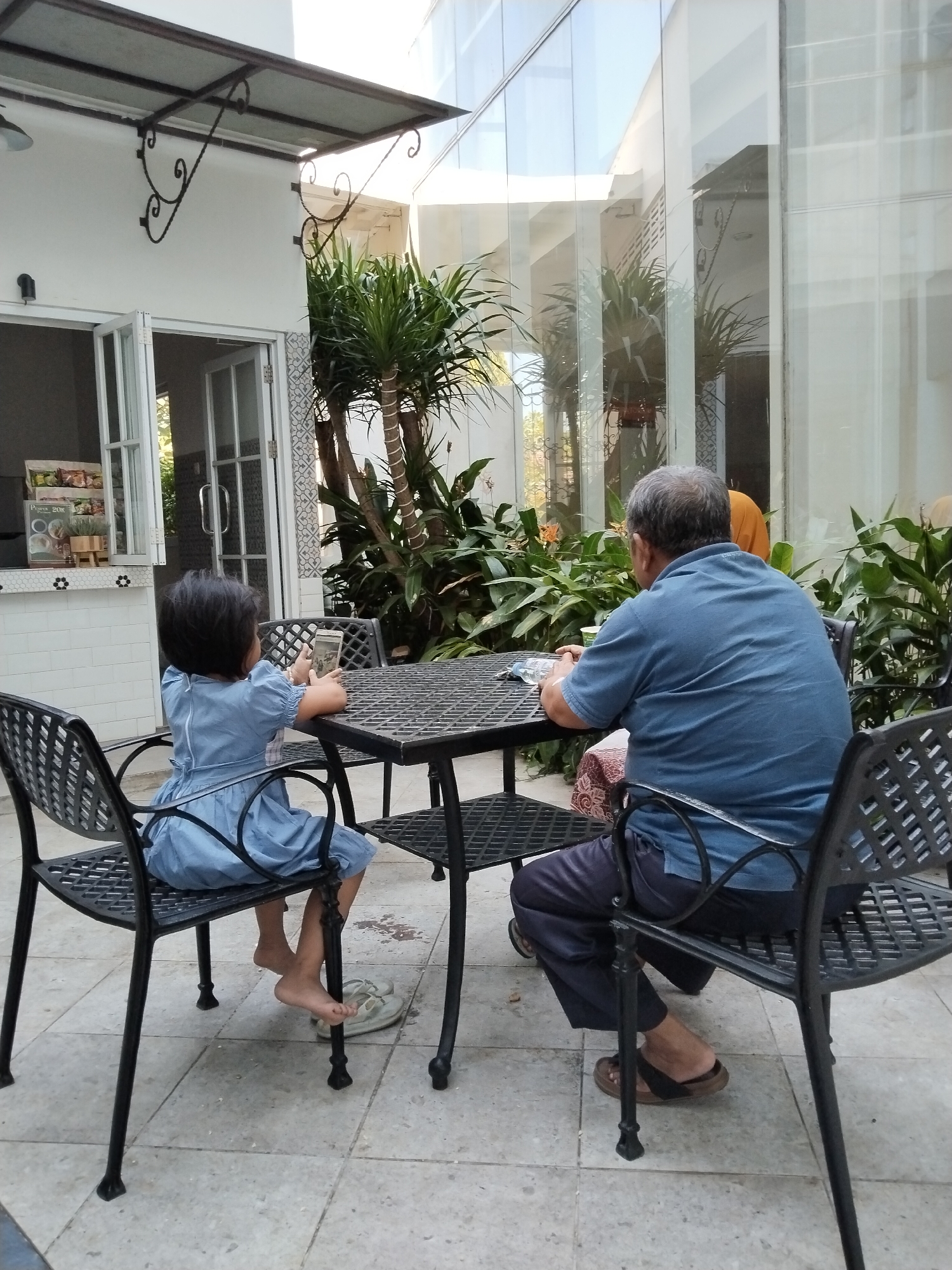 An elderly man and a young girl are seated at a table outdoors in what appears to be a courtyard or patio area. The man, wearing a blue polo shirt and dark trousers, is facing away from the camera, looking towards the girl. The girl, in a light blue dress, is also facing away from the camera, looking down at her mobile phone. Both are seated on black metal chairs at a black metal table. Behind them, a large glass-paneled building is visible, reflecting trees and the sky. The ground is tiled. In the background, there are several potted plants and part of a building with white walls and a metal awning. The scene suggests a casual dining or resting area, potentially in a cafe or restaurant in Kecamatan Pati, Indonesia.