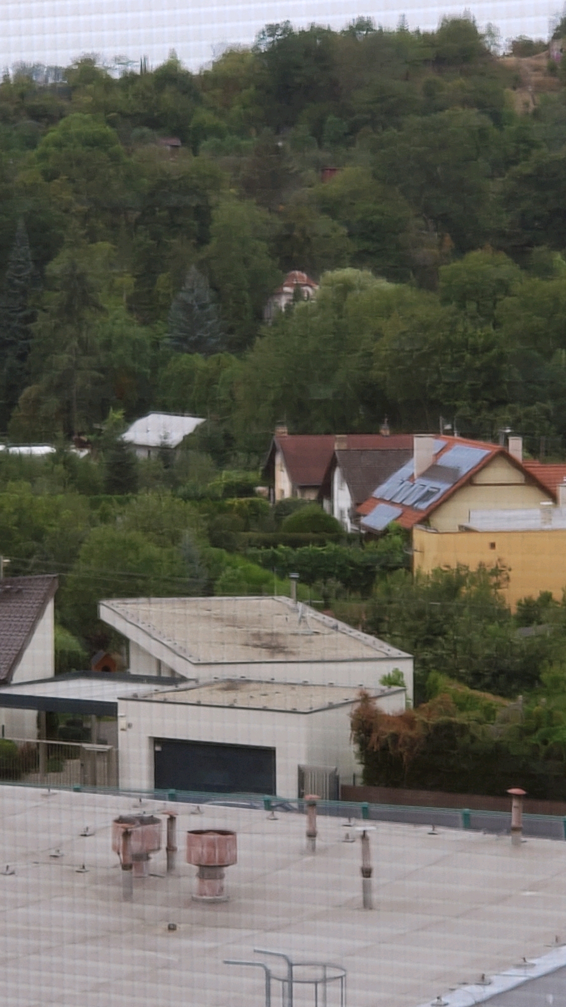 The image captures a view of residential buildings nestled on a tree-covered hillside. In the foreground, a flat, light-colored rooftop is visible, featuring several cylindrical vents with reddish-brown tops and metal bases, along with smaller metal fixtures. Behind this rooftop, a white building with a dark garage door and a covered porch area is situated. Further up the slope, several houses with pitched roofs are visible, one of which has solar panels installed on its roof. The hillside is densely wooded with a variety of trees. The overall impression is a suburban or semi-rural residential area. The location is in the city of Prešov, Slovakia.