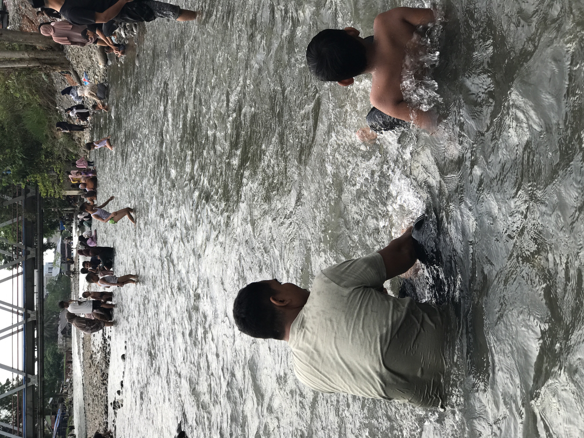 The image shows a river scene in Meurah, Indonesia, with a bridge visible on the left. Numerous people are gathered along the riverbank, some sitting or standing by the edge. Several individuals, including two boys in the foreground, are wading or swimming in the water. An adult male wearing a light-colored shirt is standing in the water, holding a dark object. The river water is flowing and slightly murky.
