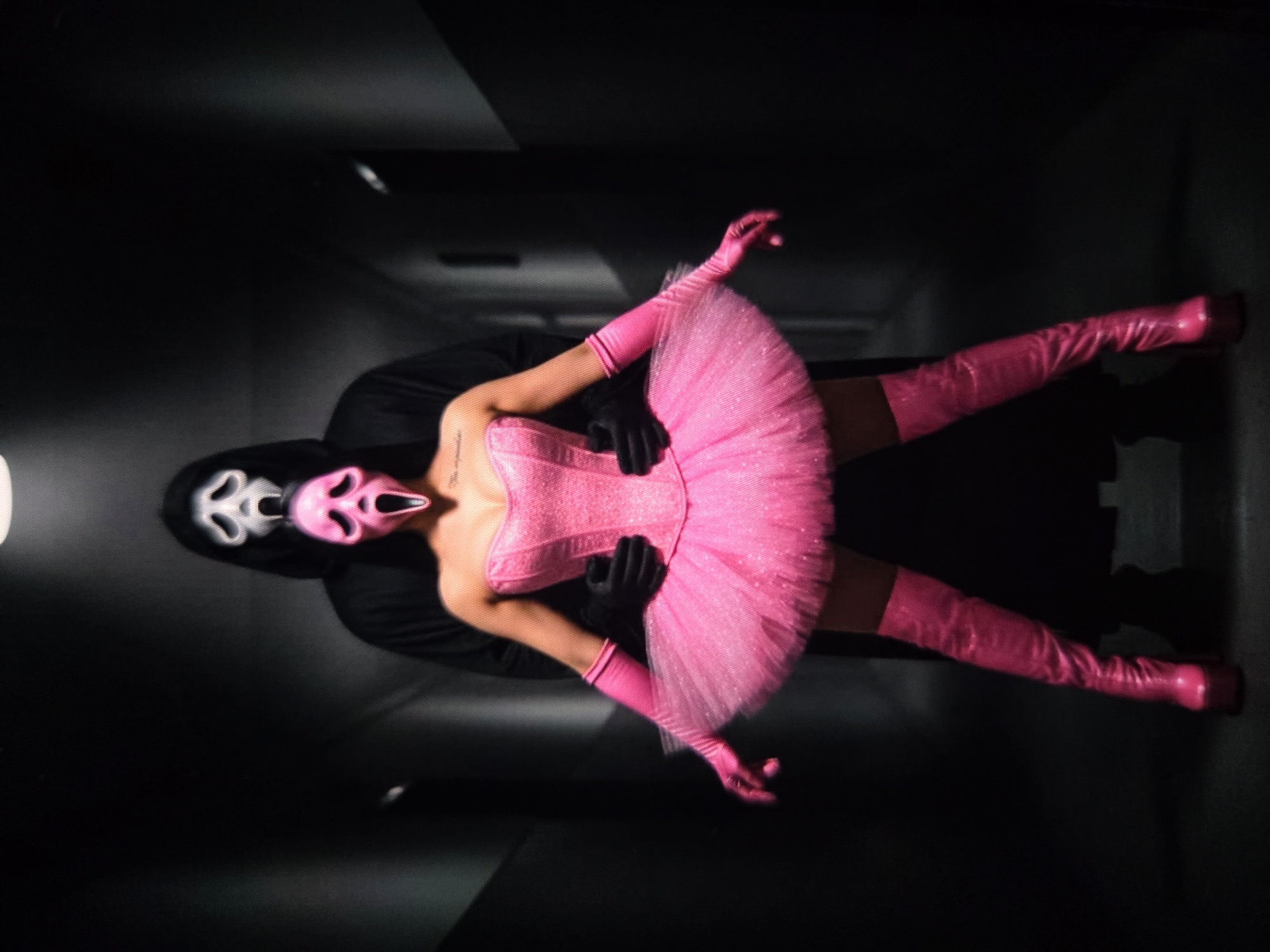 In a dimly lit, narrow corridor with dark, textured walls, two figures are positioned centrally. The foreground subject is a person wearing a bright pink, sequined strapless corset, a voluminous pink tulle tutu, matching elbow-length gloves, and knee-high platform boots. This person wears a pink Ghostface mask. Their arms are at their sides, and their legs are spread in a wide stance. Behind this figure, another person, entirely clad in black including a hooded robe and a white Ghostface mask, stands with their arms around the foreground subject's torso. The lighting emphasizes the vibrant pink attire against the dark background. There is no identifiable location in Chișinău, Moldova.