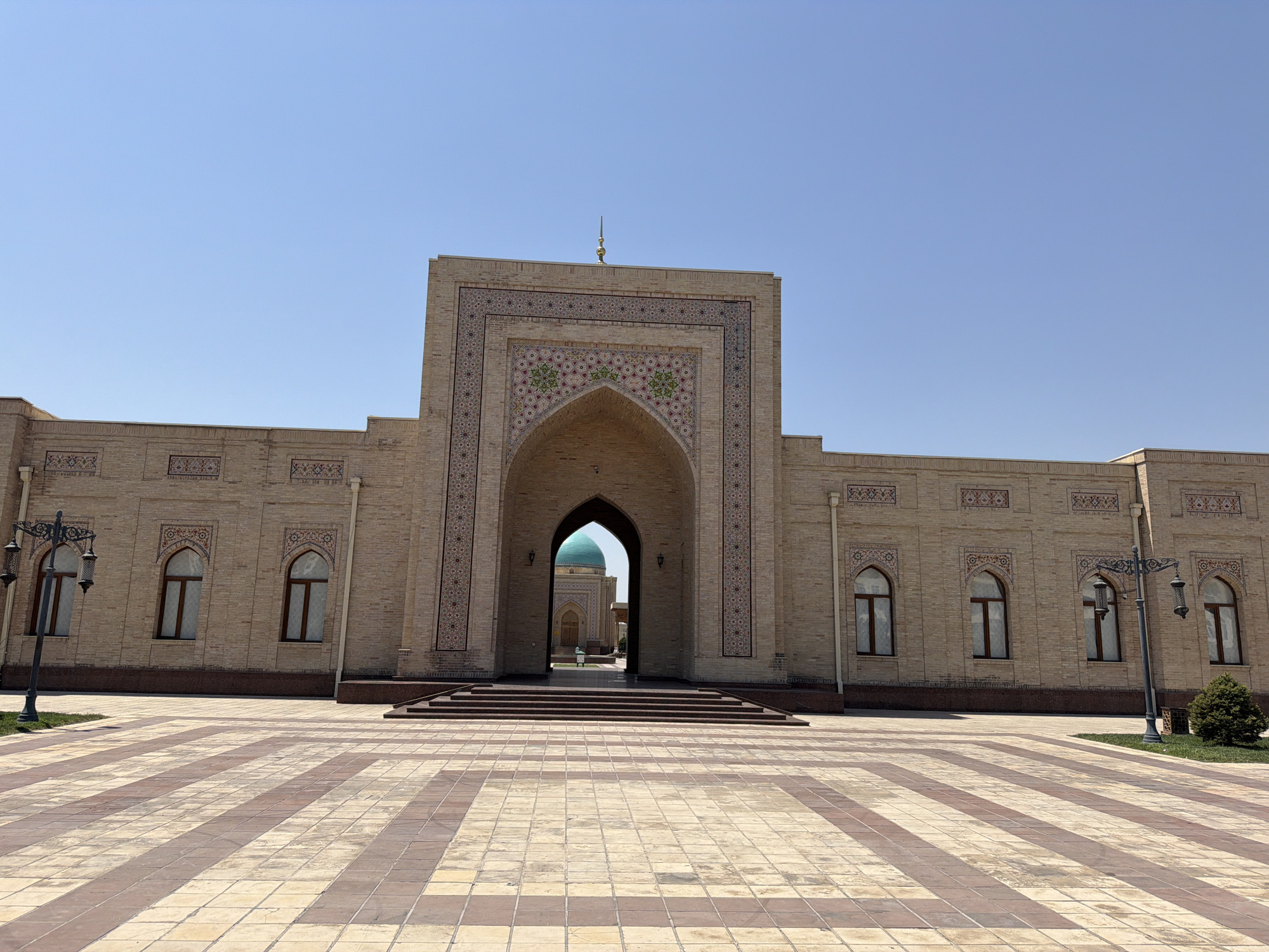 A wide-angle, eye-level, full shot captures the exterior of the Hazrati Imam Complex in Tashkent, Uzbekistan, on a clear, sunny day. The foreground features a large, paved courtyard with a striped pattern of light and dark rectangular tiles. In the mid-ground, a grand, beige brick building with intricate geometric tile work above an arched entrance dominates the center. This arched entryway leads to a view of a turquoise dome and another building in the distance. The building has a symmetrical facade with arched windows and decorative tile panels on either side of the central arch. Two ornate, black lampposts stand on the left and right sides of the courtyard, flanking the main building. A small, manicured evergreen bush is visible on the right side of the image. The sky is a clear, bright blue with no clouds. The scene is devoid of any people or animals, presenting a serene and architectural view of the historical complex.