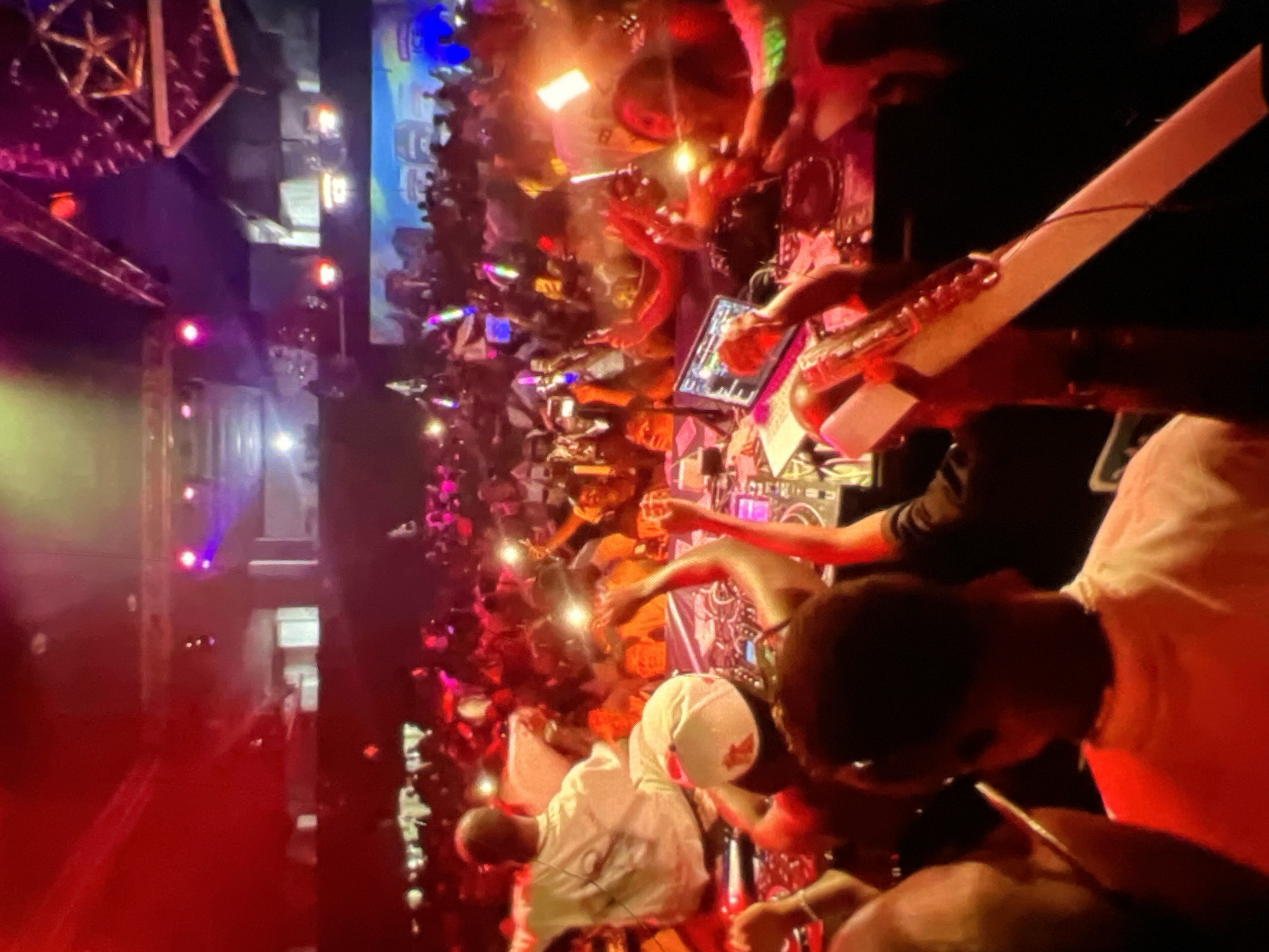 The image depicts a lively scene at a nightclub in Lagos, Nigeria, illuminated by vibrant red and purple lights. A large crowd of people are visible, many holding up their phones with their lights on, creating a festive atmosphere. In the foreground, several individuals are gathered around DJ equipment, suggesting a performance or party in progress. One person, wearing a white shirt and a cap, is facing away from the camera, while another person in a white t-shirt is partially visible. A laptop is open on a DJ booth, and various electronic devices are present. The overall impression is of an energetic social gathering, characteristic of the nightlife in Lagos.