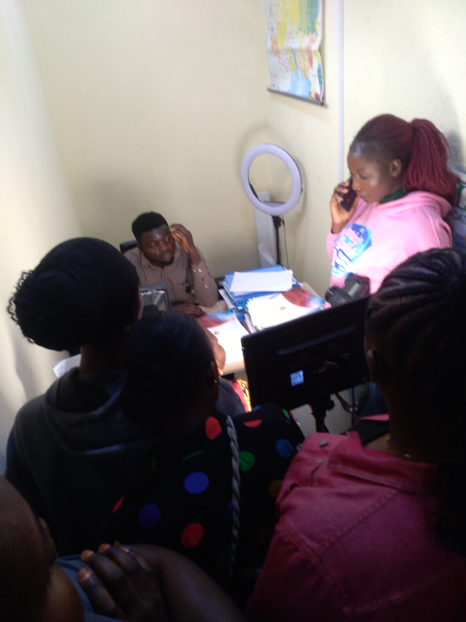 The image captures an indoor gathering of several individuals, likely in an office or educational setting in Jos, Nigeria. The scene is centered around a desk with multiple people present, suggesting a collaborative or instructional activity.
In the right foreground, a woman with red hair styled in a ponytail, wearing a pink hoodie with a graphic design, is actively speaking on a mobile phone, holding it to her right ear. To her left, behind a desk, a man in a brown long-sleeved shirt is seated, looking down at a screen or papers on the desk, with his hand near his face.
The desk area is equipped with several items: a bright white ring light on a stand is prominent next to the woman on the phone, indicating potential use for video calls, photography, or content creation. A black computer monitor faces away from the camera, and a dark-colored DSLR-type camera rests on the desk. Various papers, books, and folders are also visible on the desk surface.
Several other individuals are partially visible, mostly from behind, observing or participating in the activity. One person in the left foreground wears a dark hoodie, while another wears a dark top with colorful polka dots (red, blue, green). A third person in the right foreground wears a pink collared shirt.
On the light-colored wall behind the desk, a colorful world map is pinned, adding to the impression of a learning or globally-oriented environment. The lighting in the room appears to be from artificial sources, possibly including the ring light itself, as well as overhead lights, creating an evenly lit but somewhat warm ambiance. The overall atmosphere suggests focused attention and engagement among the group. No specific time of day or outdoor weather conditions can be discerned. Text on a sign behind the woman in pink is partially visible, starting with "DATI...".