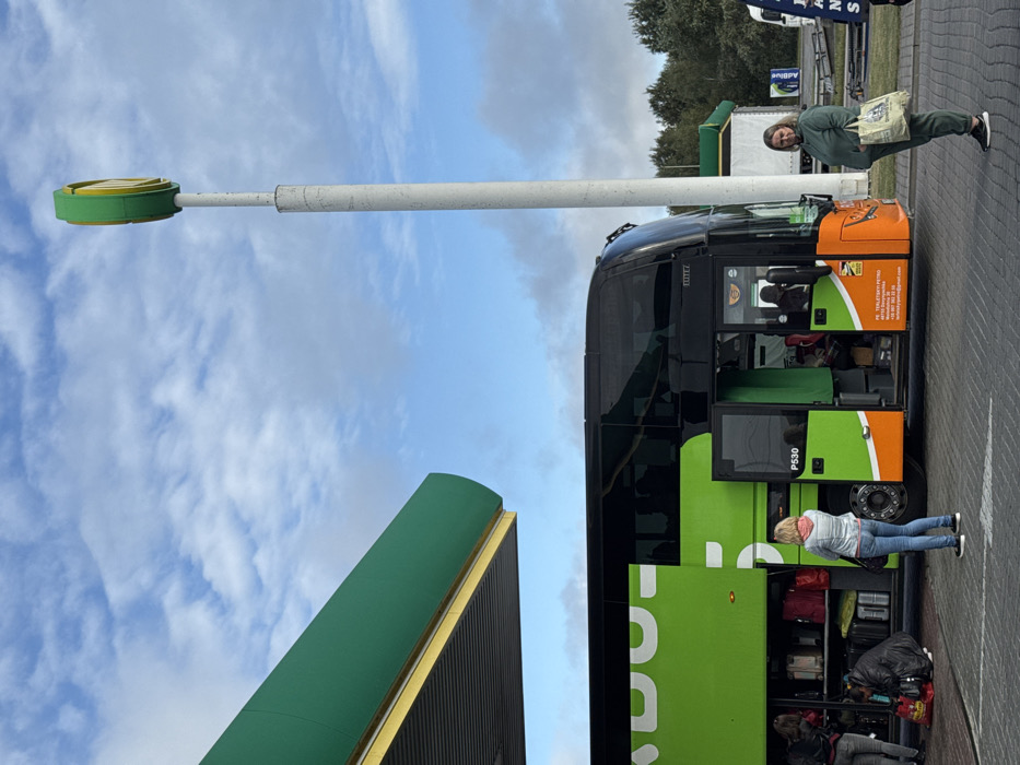 The image depicts an outdoor scene at a service stop in Starovoitove, Ukraine, under a partly cloudy sky. A large, black and green FlixBus double-decker coach, identified by "P530" and displaying "PS TERLETSKY PETRO" with contact information on its lower front panel, is centrally positioned. Its front passenger door is open, revealing an individual seated inside. A side luggage compartment door is also open, exposing multiple suitcases, bags, and red parcels. Two individuals are located near this compartment; one kneels, actively handling luggage, while another stands next to them. A third woman, with blonde hair and wearing a light blue denim jacket over a pink top and blue jeans, stands facing the bus, observing the luggage area. To the right of the bus, a fourth woman, dressed in a matching olive-green tracksuit, stands facing the viewer, holding a light-colored canvas bag with blue lettering in her left hand. The immediate environment includes a green overhead canopy covering part of the scene and a tall white pole topped with a green and yellow circular sign. The ground is paved, and trees are visible in the distant background.