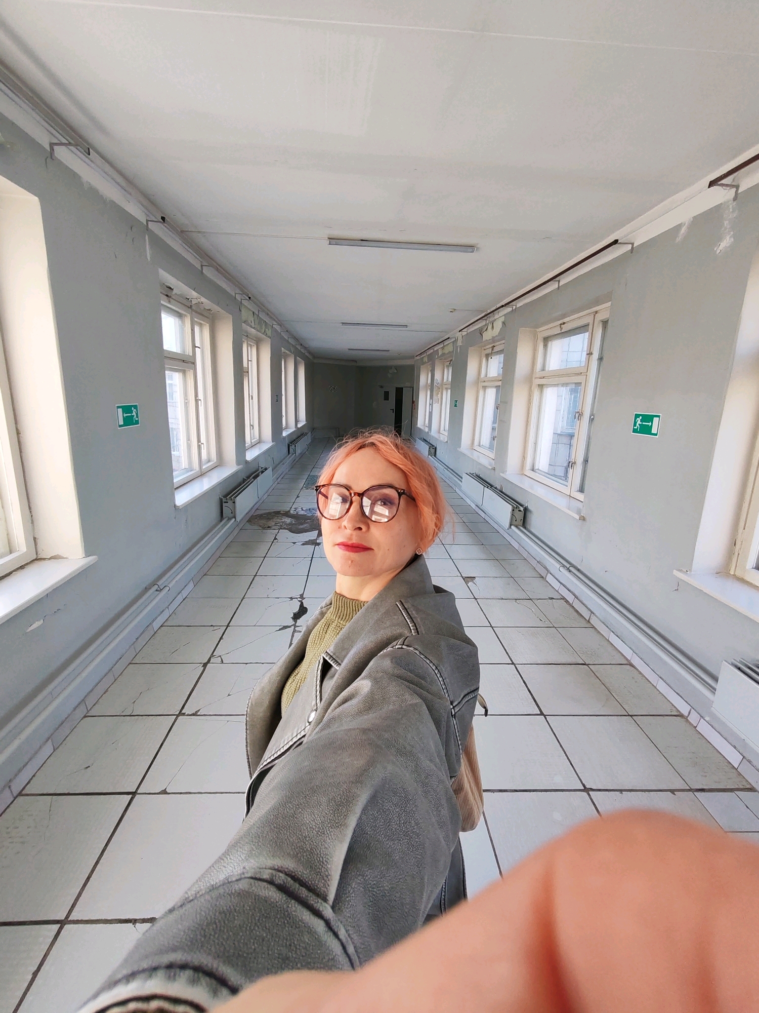 A woman with pink hair and glasses stands in a long, sterile hallway, taking a selfie. The hallway is lined with identical windows on either side, revealing a city scene outside that appears to be Chaikovsky, Russia, based on context. The floor is tiled with a grid pattern, and the walls are painted a pale gray. Two green emergency exit signs are visible on the walls. The overall impression is one of an institutional or abandoned building. The woman is wearing a gray jacket over a green sweater.